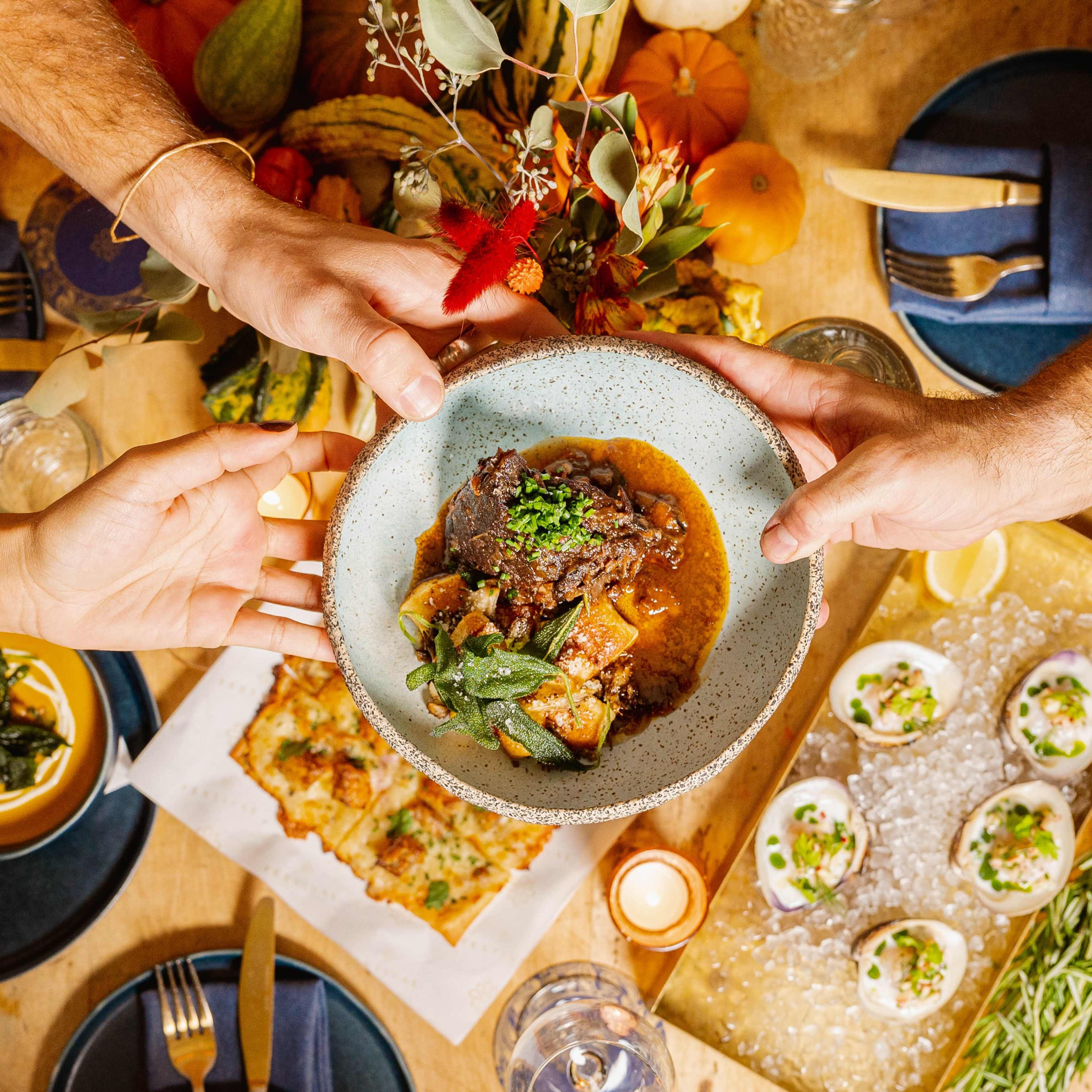 Two hands reach for a plate of stew at a festive table filled with various dishes and autumn decorations.