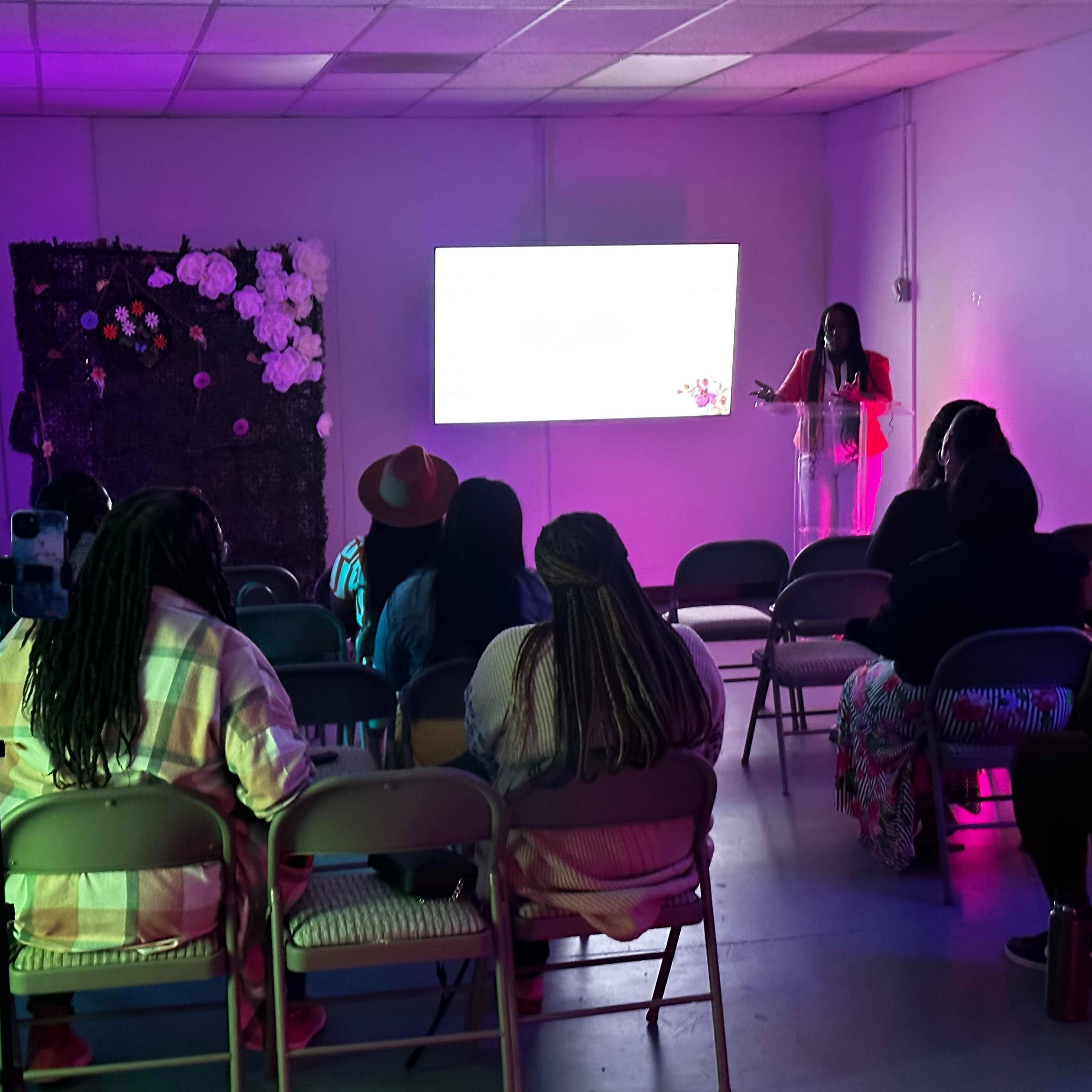 A speaker addresses an audience seated in a brightly lit room decorated with flowers on a screen.
