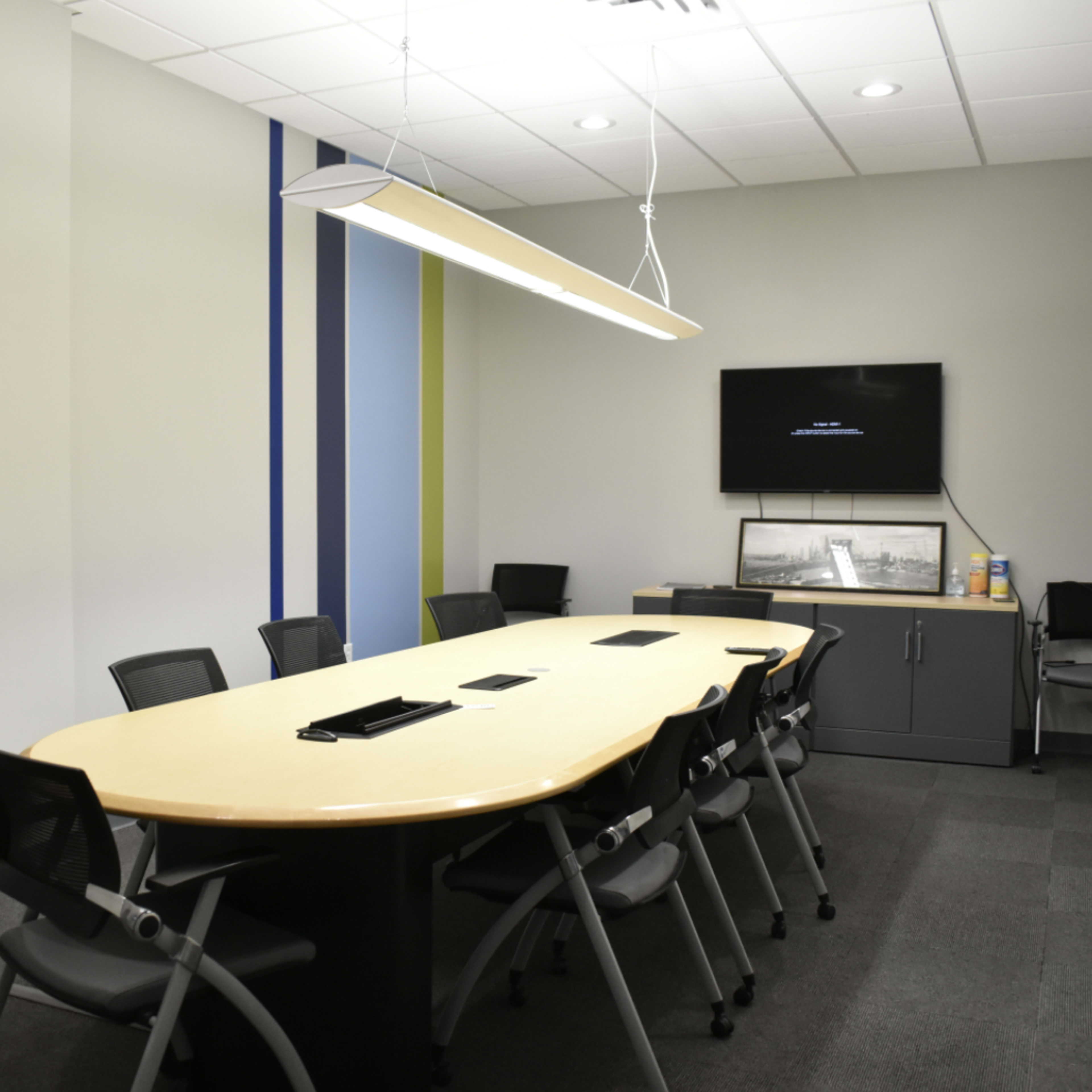 A modern conference room features a large oval table surrounded by black chairs, with a television mounted on the wall and colored stripes decorating the walls.