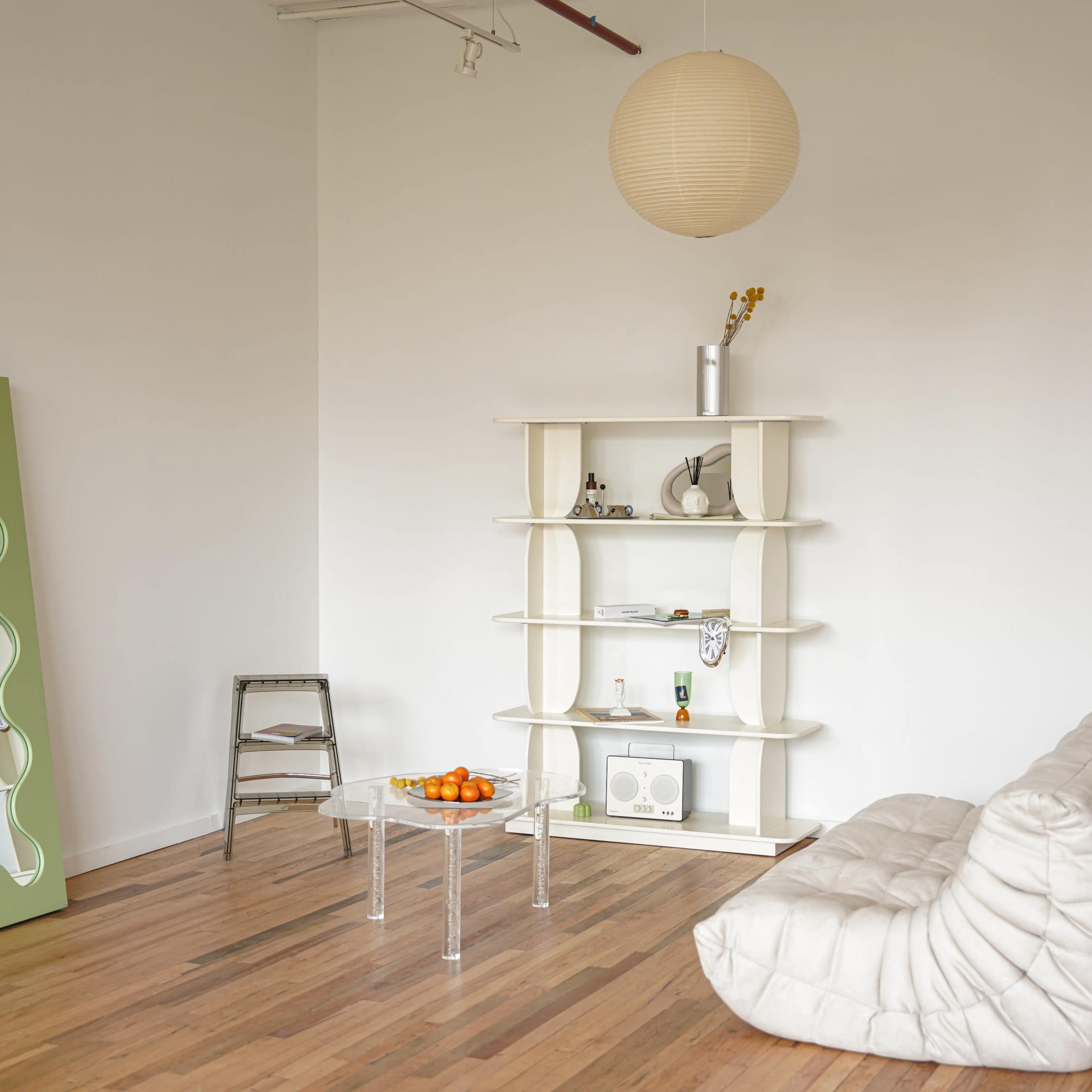 A minimalist room features a white tufted sofa, a green mirror, a shelf with decorative items, and a bowl of oranges on a clear table.