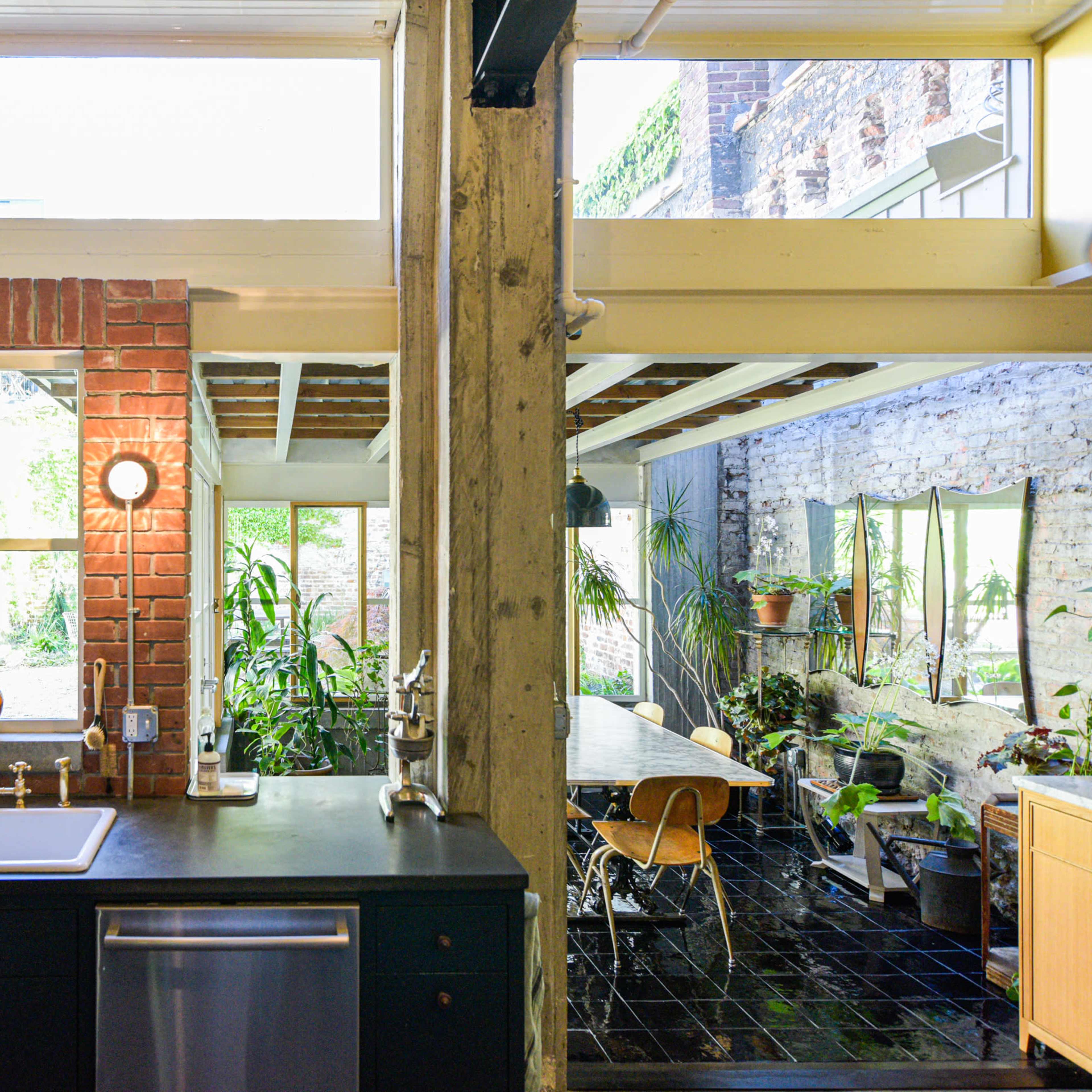 A modern kitchen with a black countertop and stainless steel appliances, adjacent to a sunlit dining area featuring greenery and large windows.