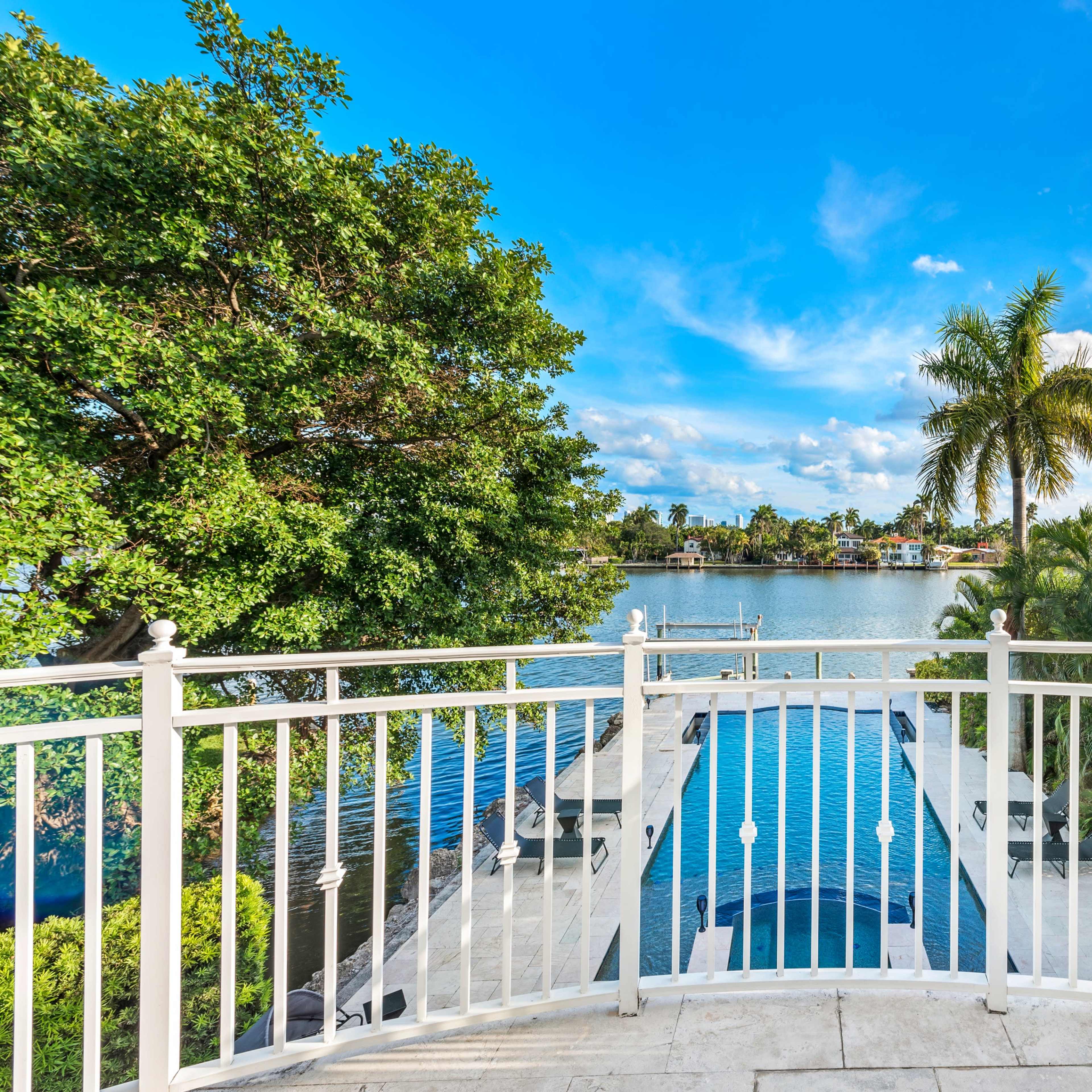 A waterfront view from a balcony overlooking a pool and dock surrounded by lush greenery and palm trees.