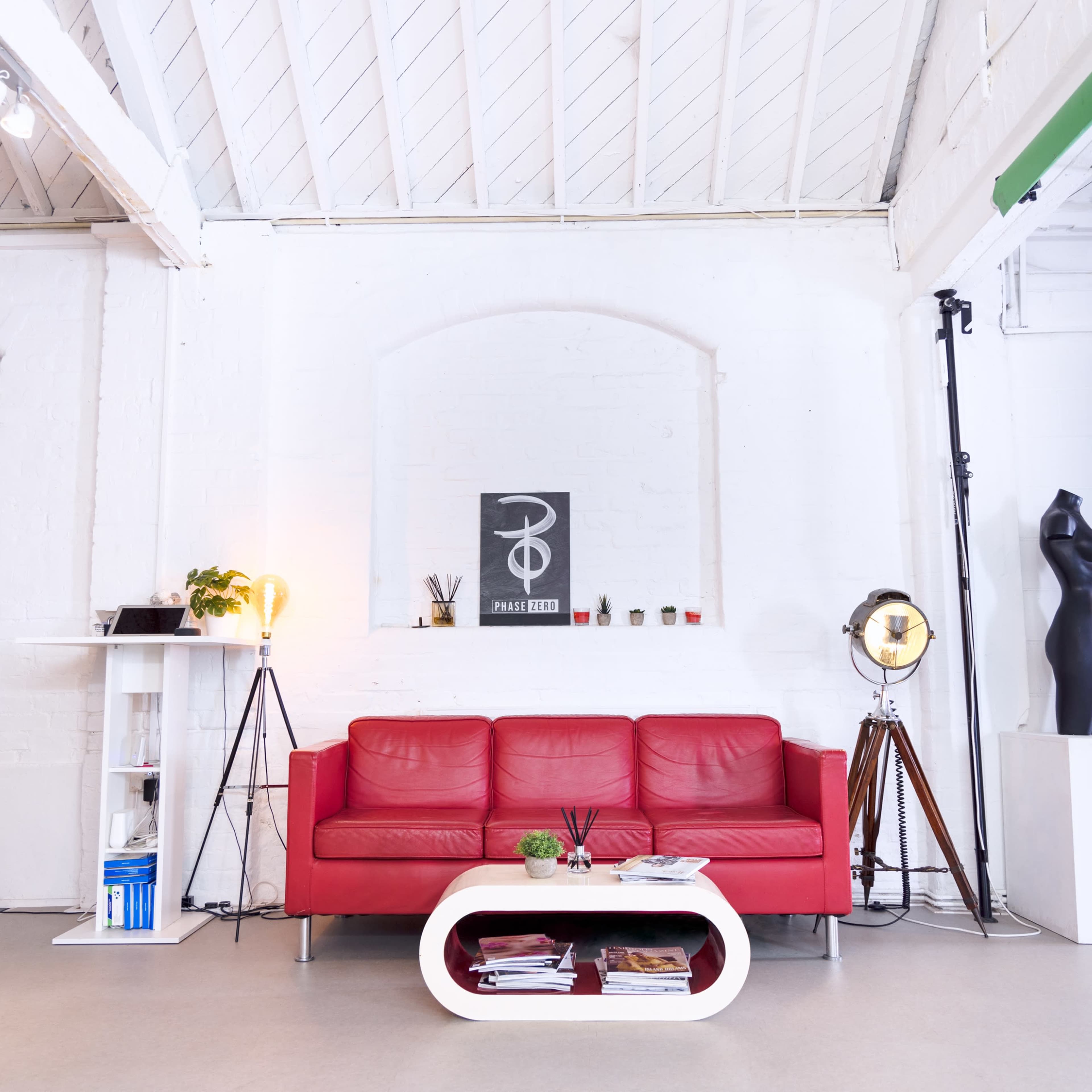 A red sofa is positioned in a bright, open room featuring white walls and a high ceiling, with a decorative stand and an artwork on the wall.