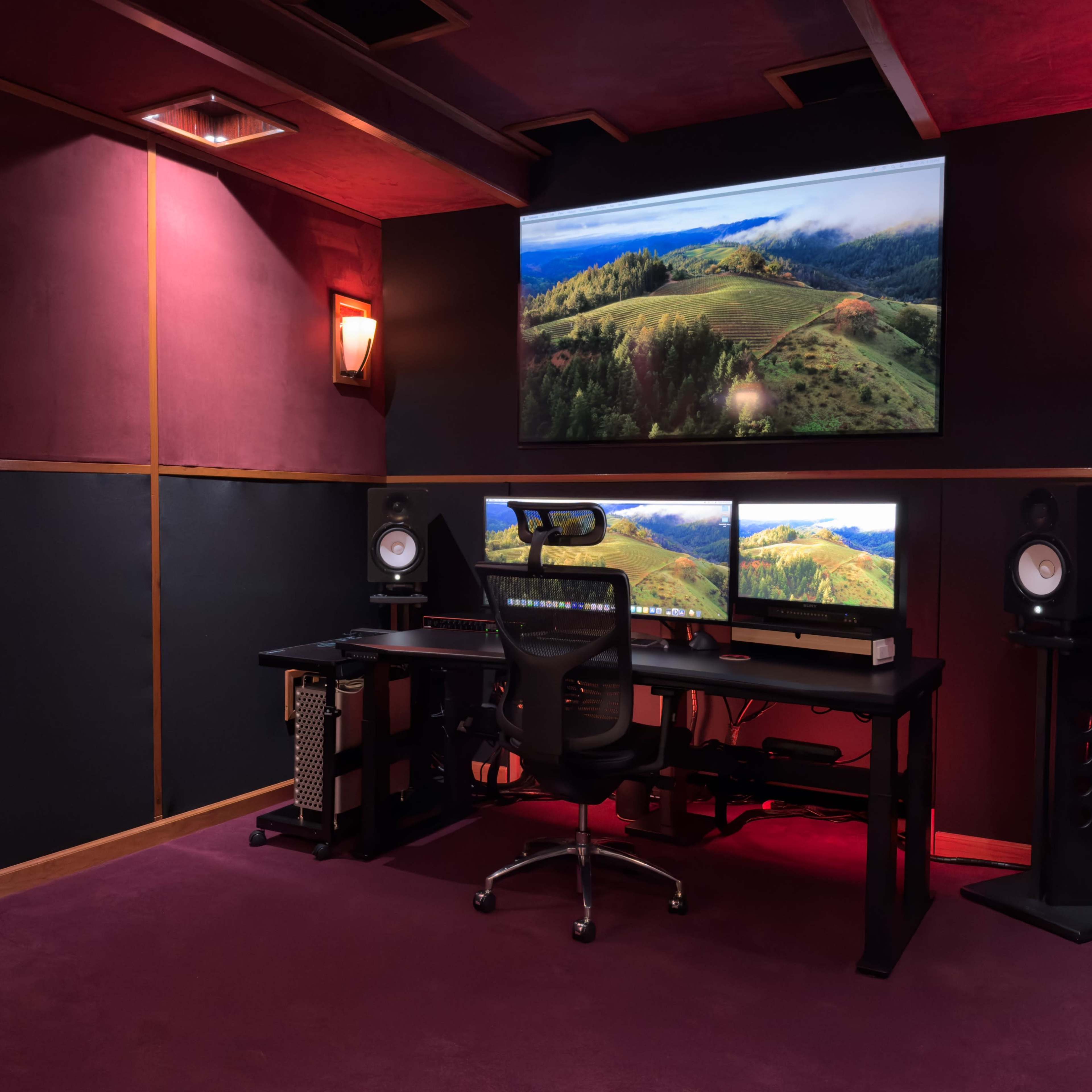 A sound studio featuring a desk with two computer monitors, a chair, and large speakers, all situated against dark walls with a large screen displaying a landscape.