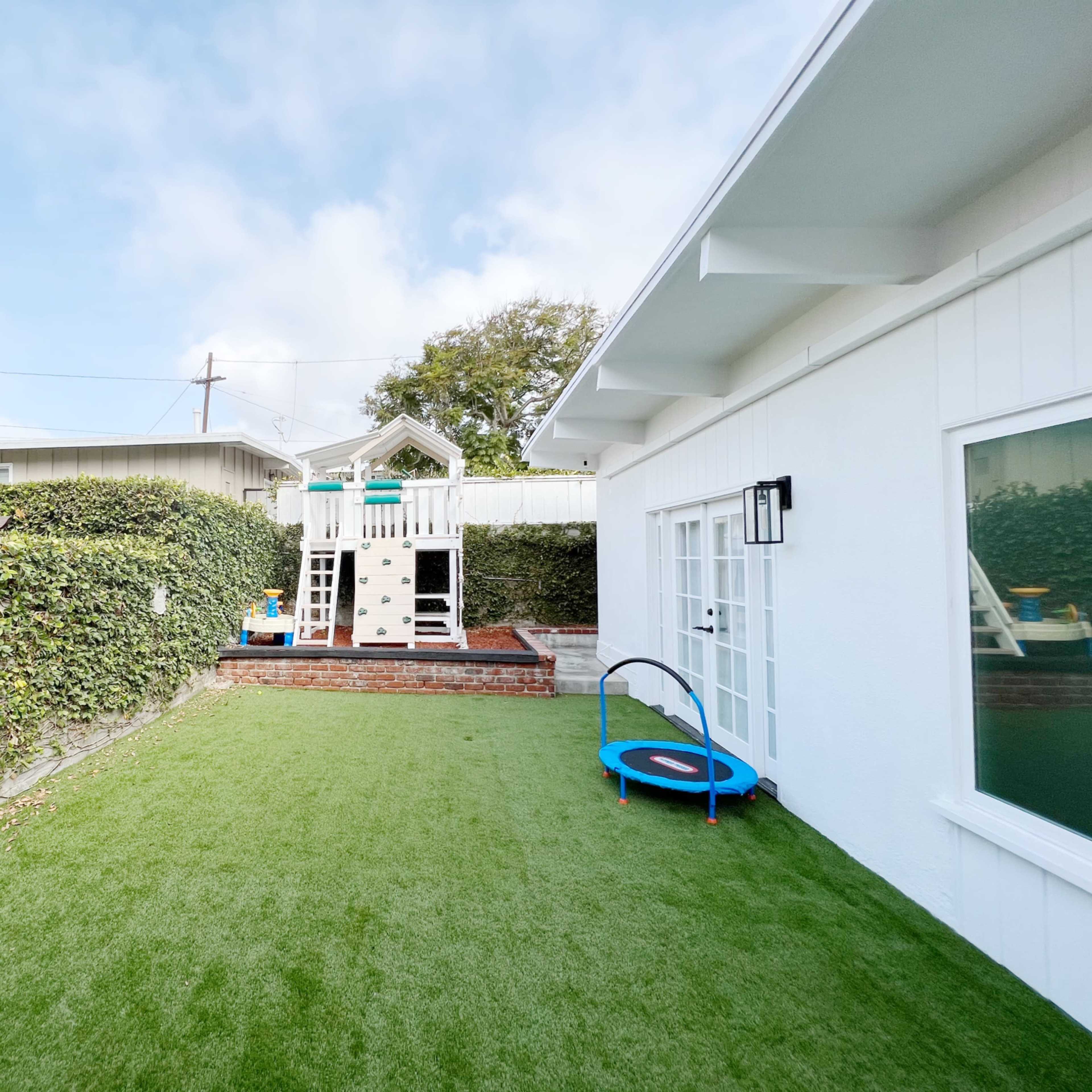 A backyard with artificial turf, a trampoline, and a play structure surrounded by a low wall and greenery.