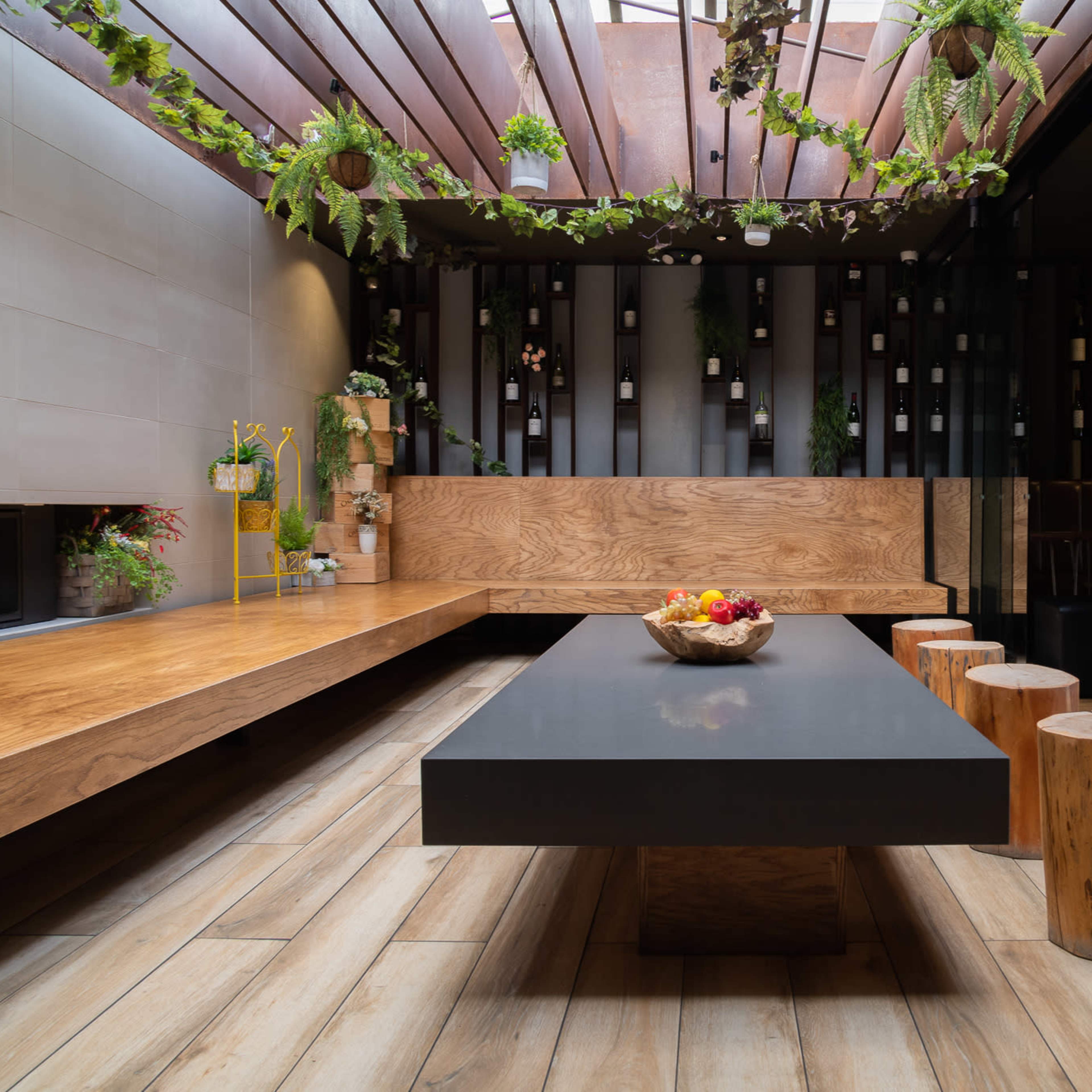 A modern interior space featuring a long black table surrounded by wooden stools and a bank of potted plants overhead.