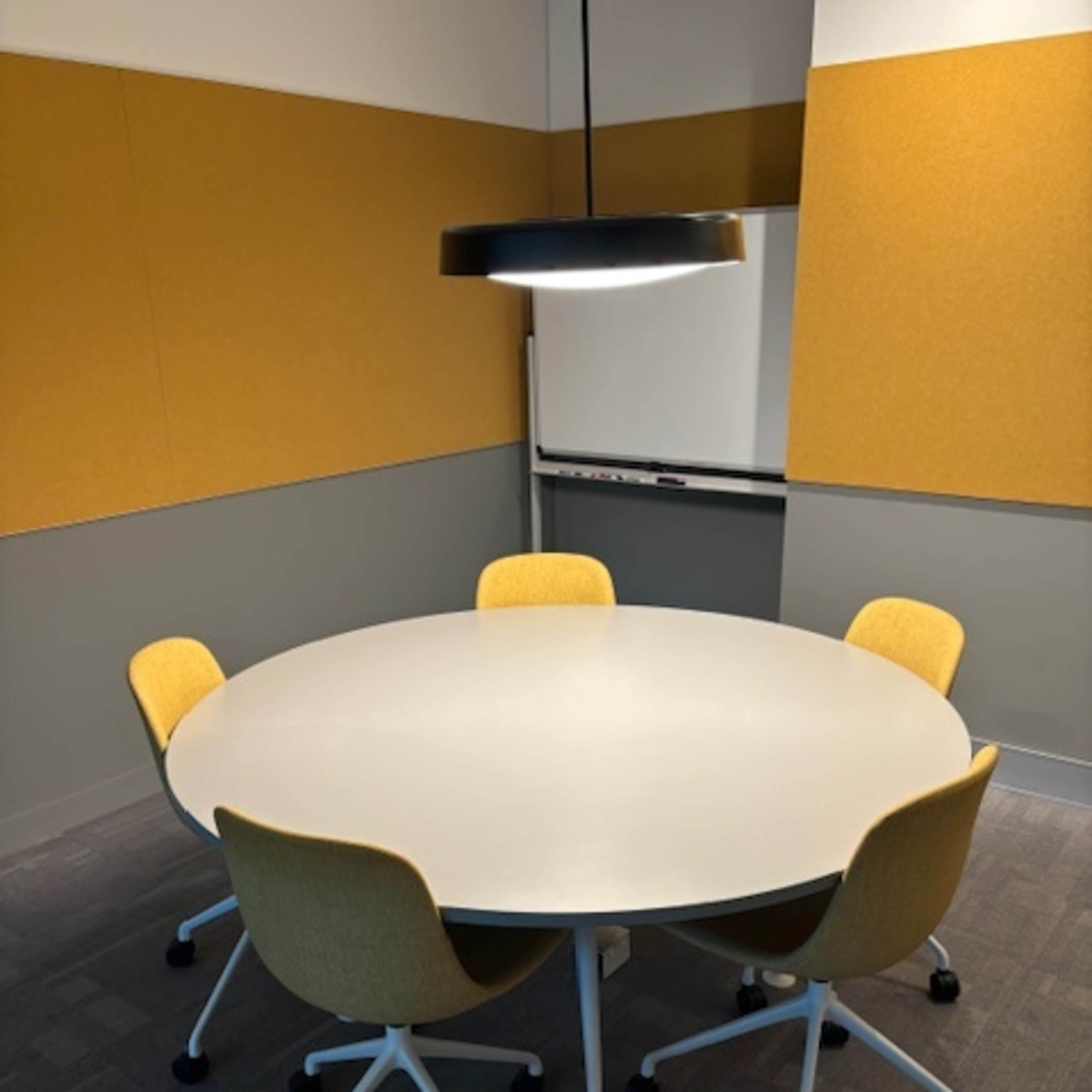 A round table with four yellow chairs is set in a conference room featuring grey and yellow walls.