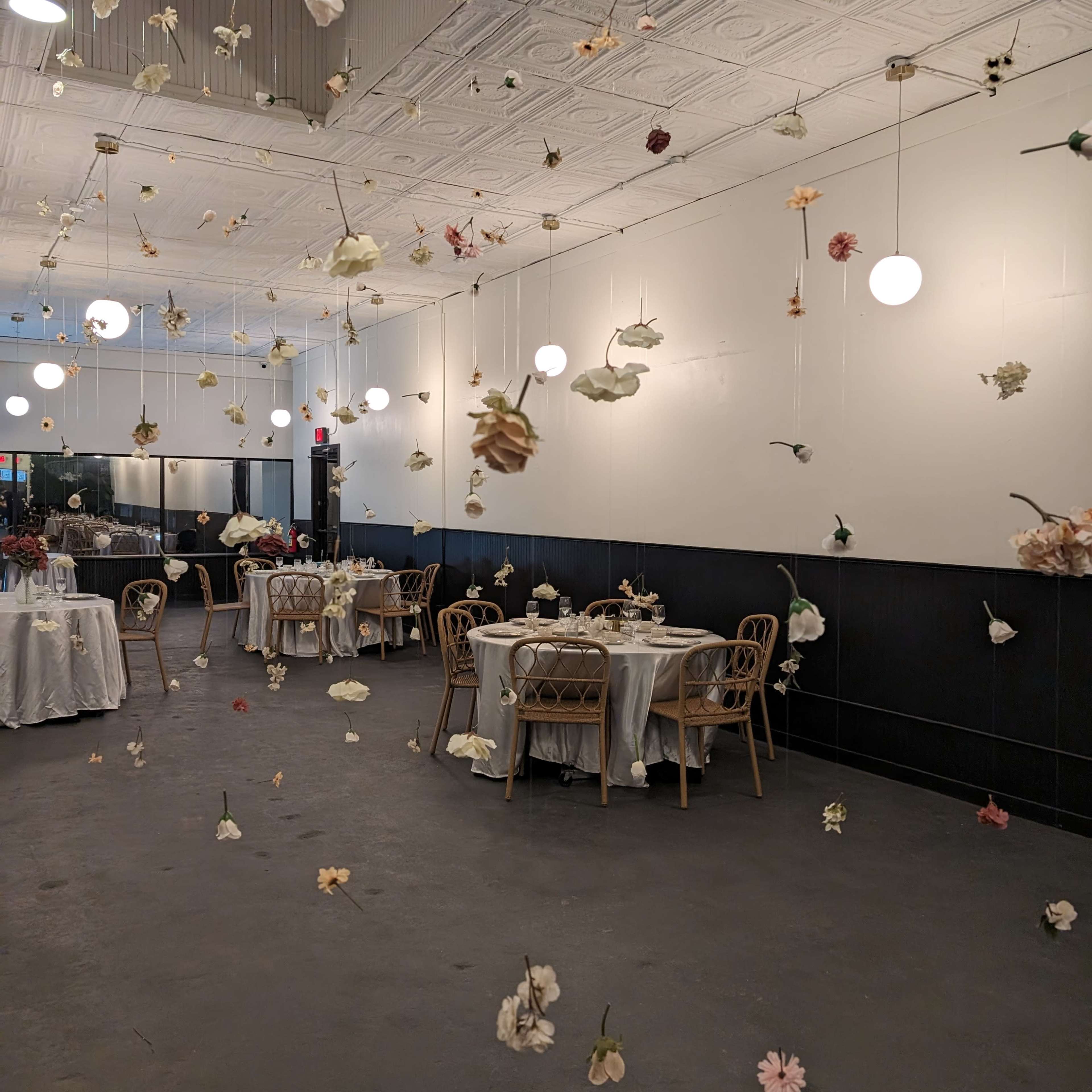 A dining area with tables set for a meal, decorated with hanging flowers and illuminated by round pendant lights.