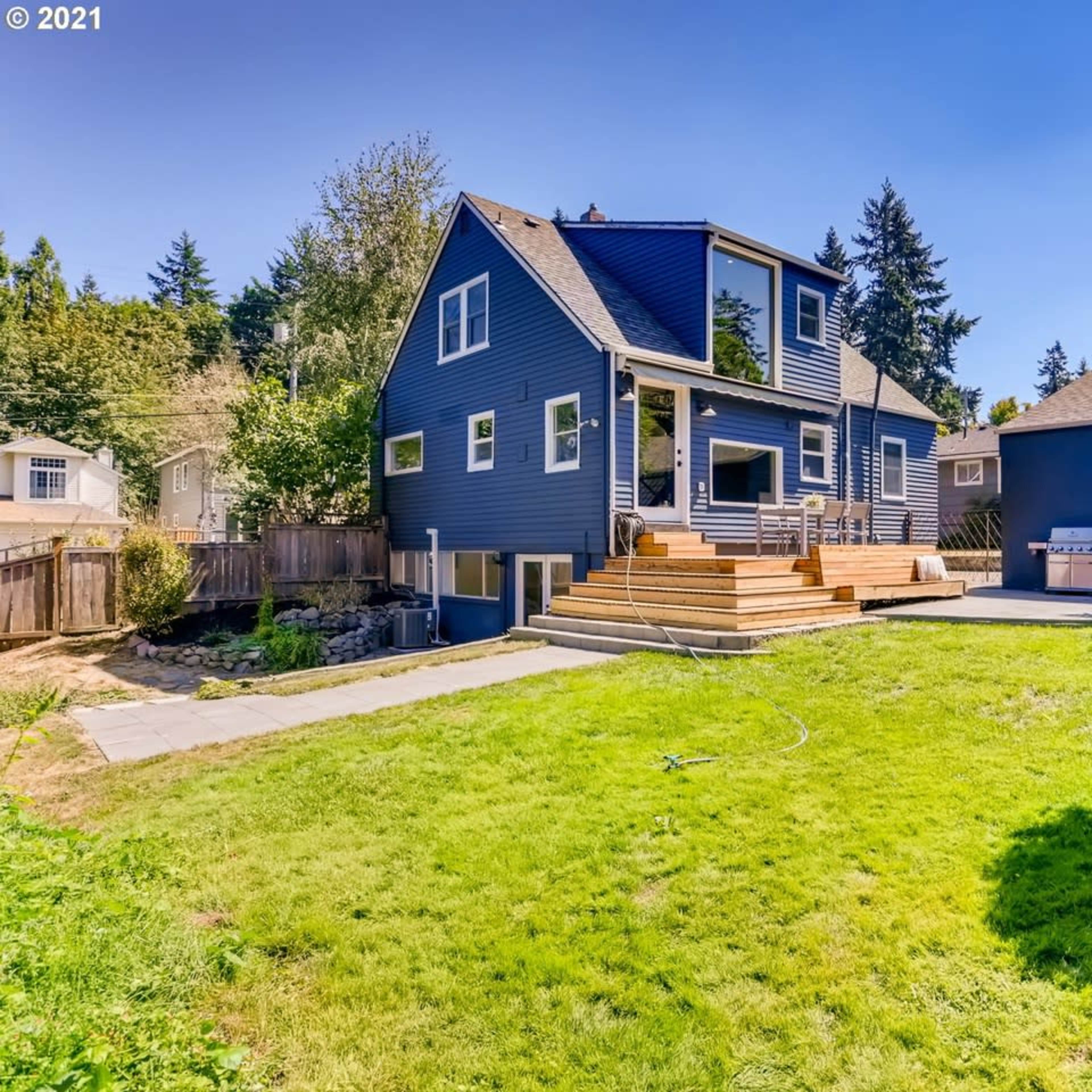 A blue two-story house in a yard with a wooden deck, surrounded by trees and greenery.