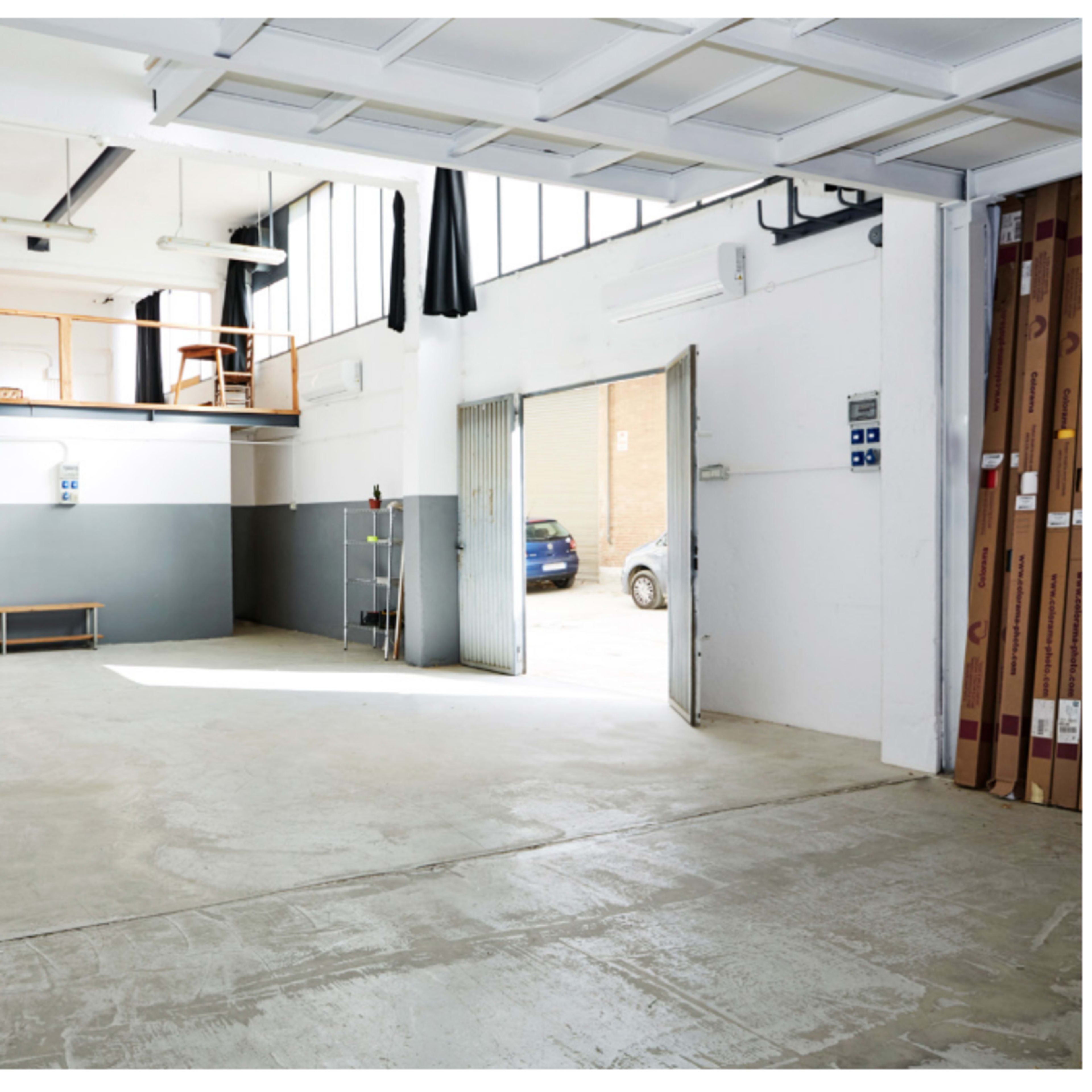 A spacious, well-lit industrial interior with a loft area and an open entrance, featuring concrete flooring and large windows.