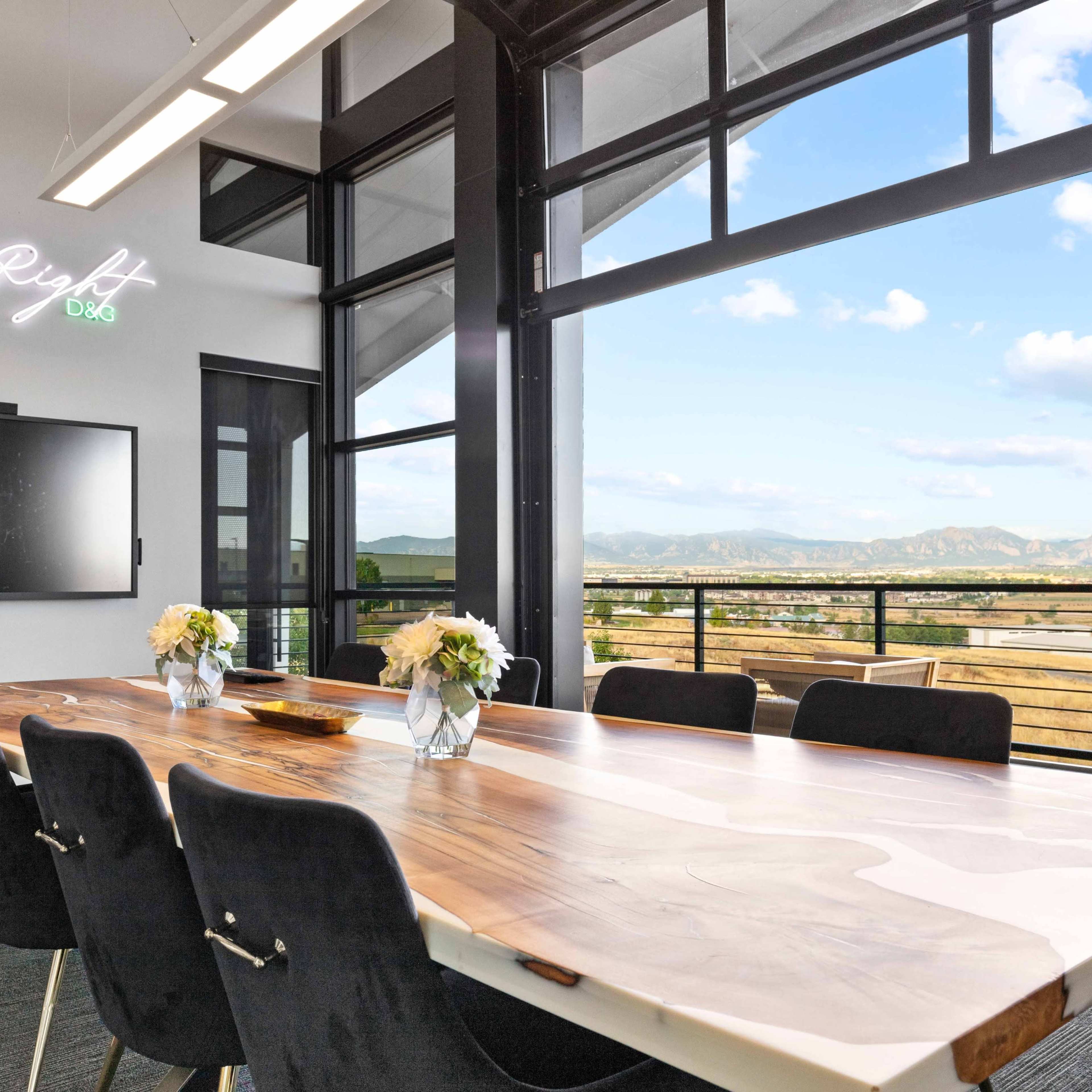 A modern conference room features a long wooden table, black chairs, and large windows with a view of mountains and the sky.