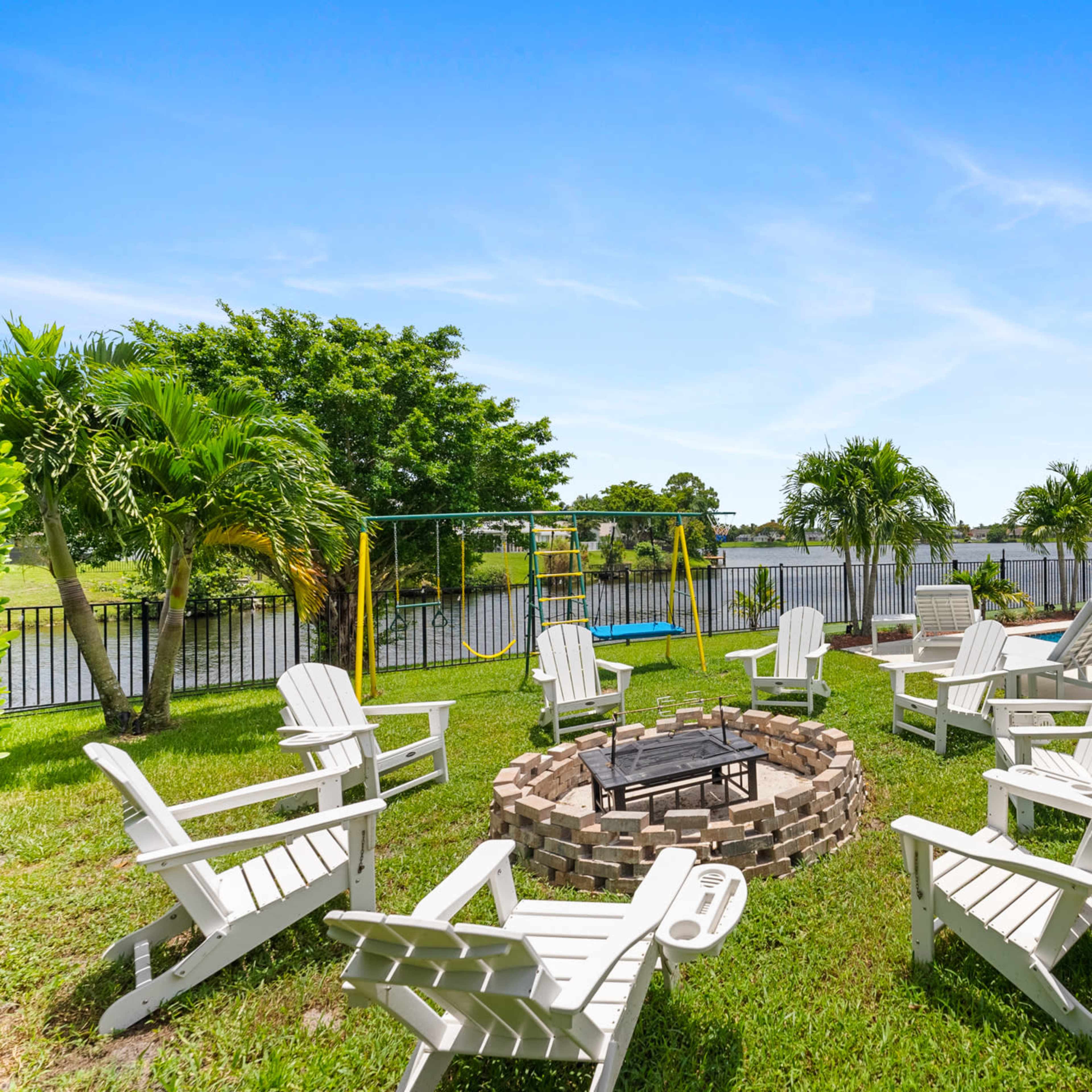 A circular stone fire pit surrounded by white Adirondack chairs is set on the grass next to a swimming pool and a waterfront view.