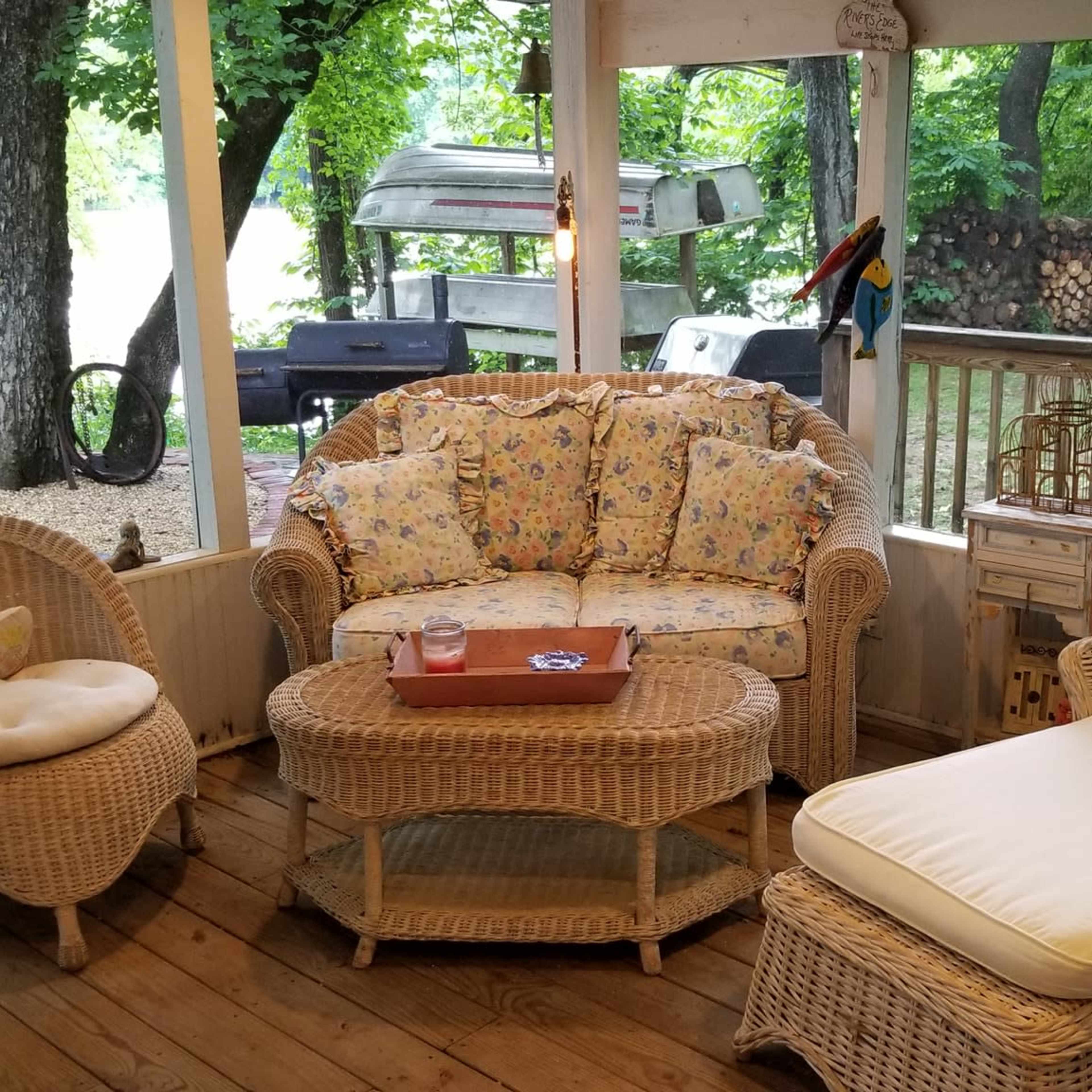 A screened-in porch featuring woven furniture, including a couch, a chair, and a chaise lounge, arranged on a wooden floor with a view of greenery outside.