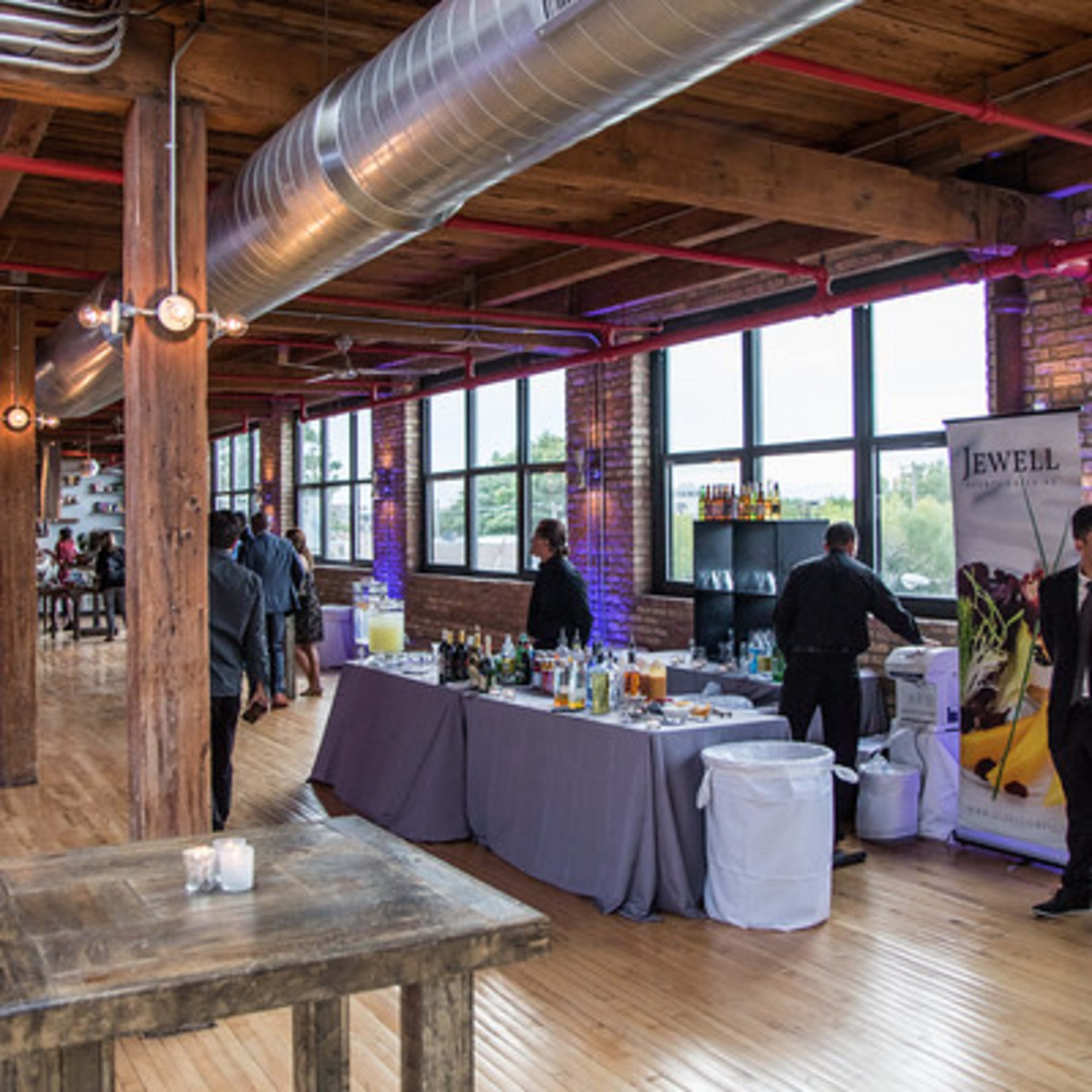 A spacious event venue with wooden beams, large windows, and a bar setup featuring various drinks and decorations.