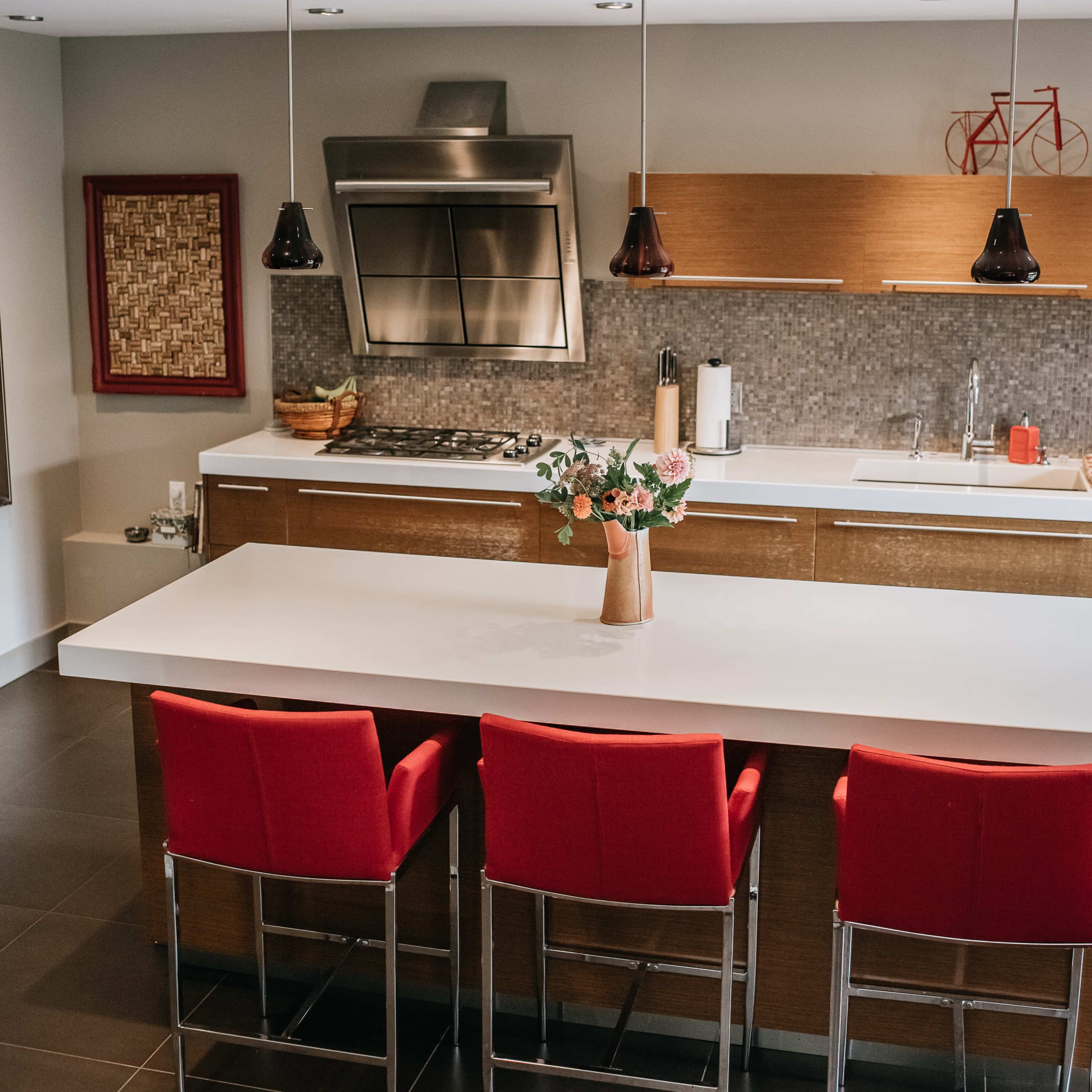 A modern kitchen features a central island with three red bar stools, stainless steel appliances, and a decorative vase of flowers.