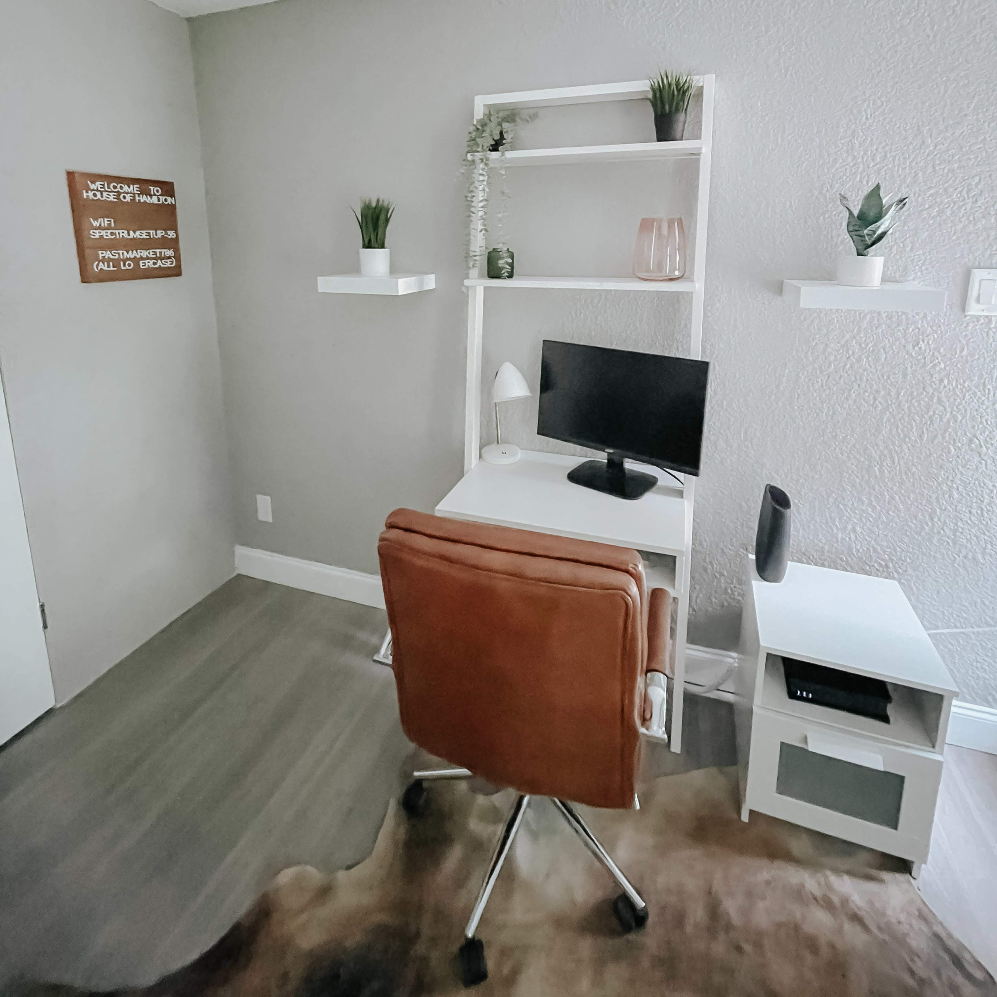 A minimalist home office featuring a desk with a computer, a brown chair, plants on shelves, and a sign on the wall.