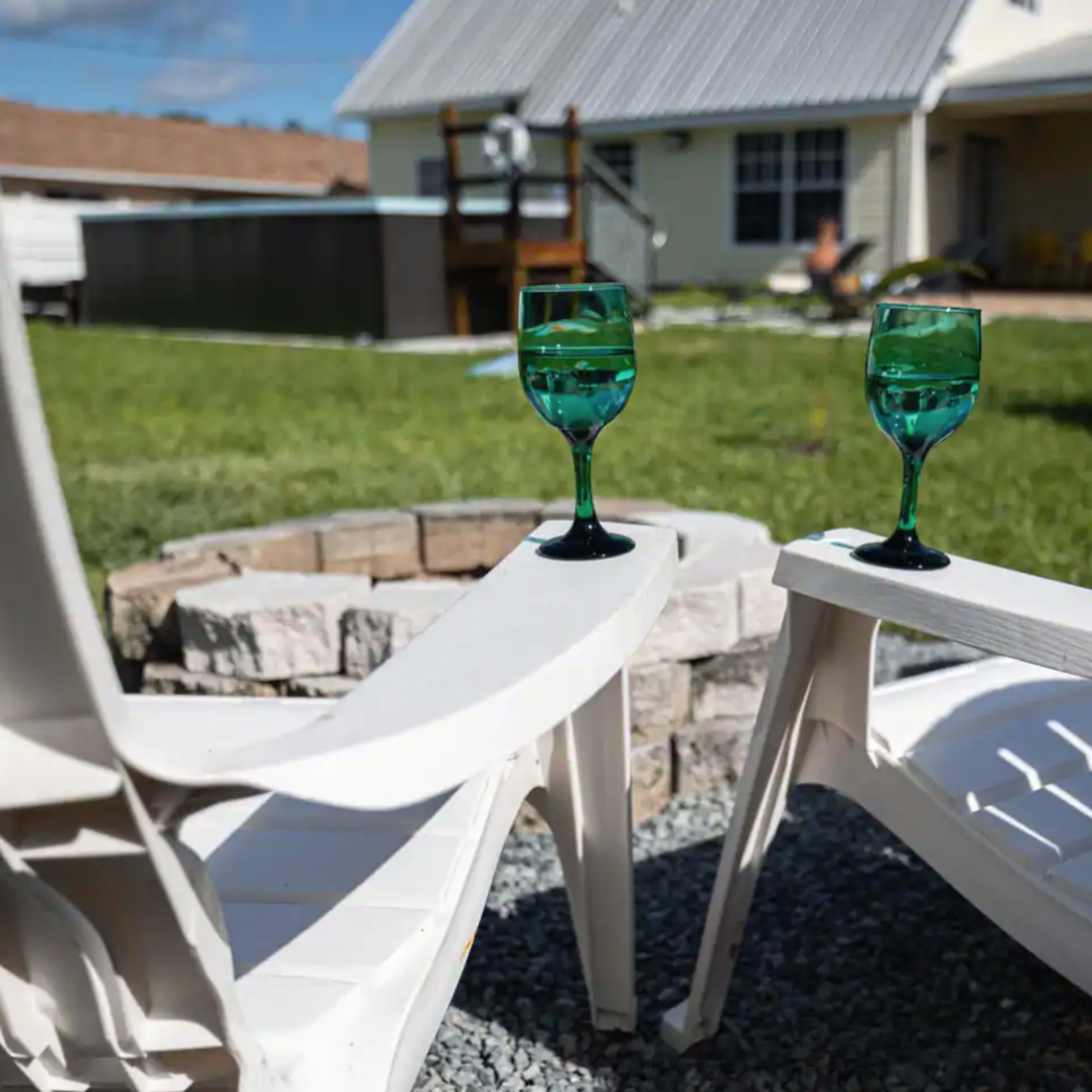Two turquoise glasses sit on a white plastic table between two lounge chairs beside a fire pit in a backyard.