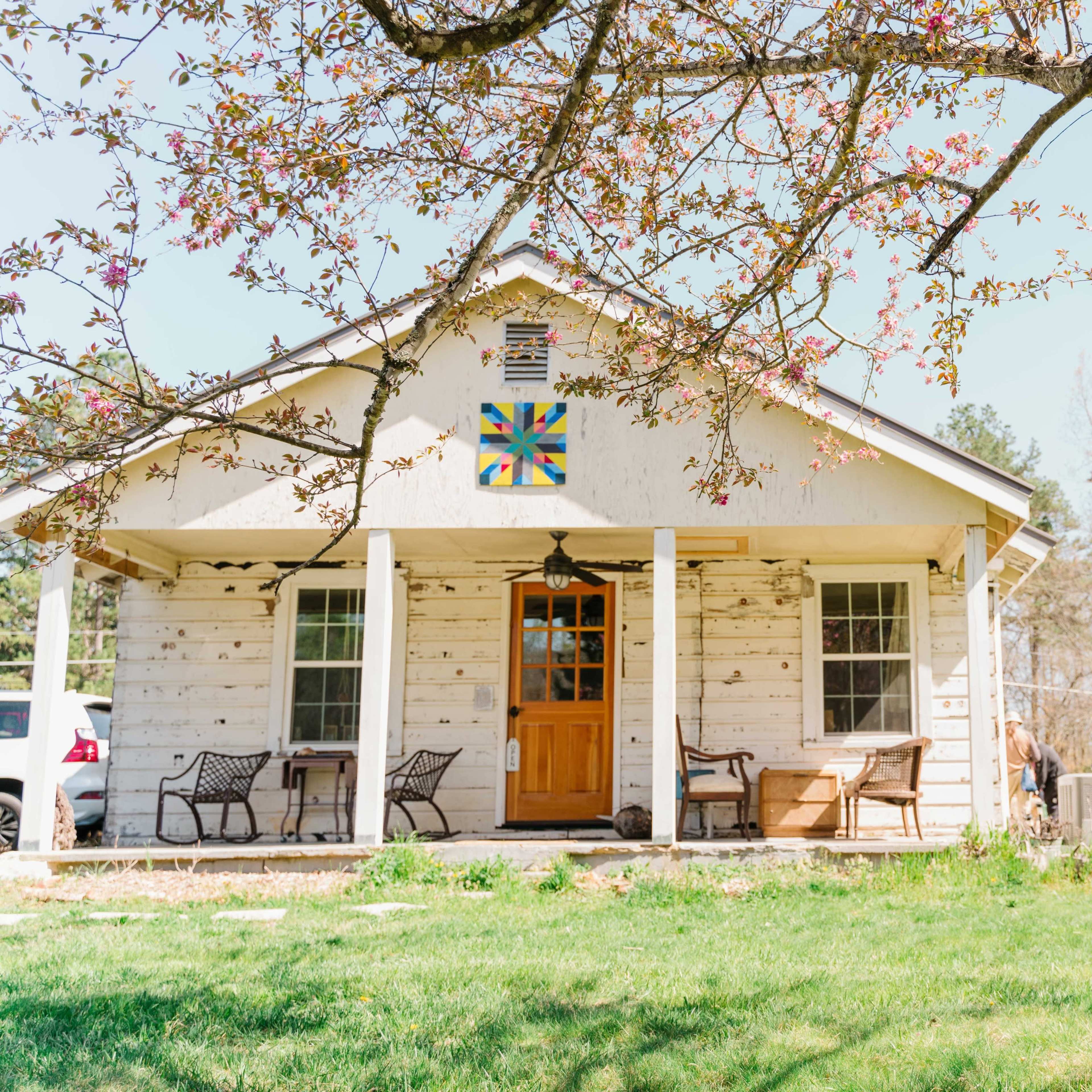 A rustic house with a flower-patterned quilt square on the front, surrounded by a grassy yard and several people gathered nearby.