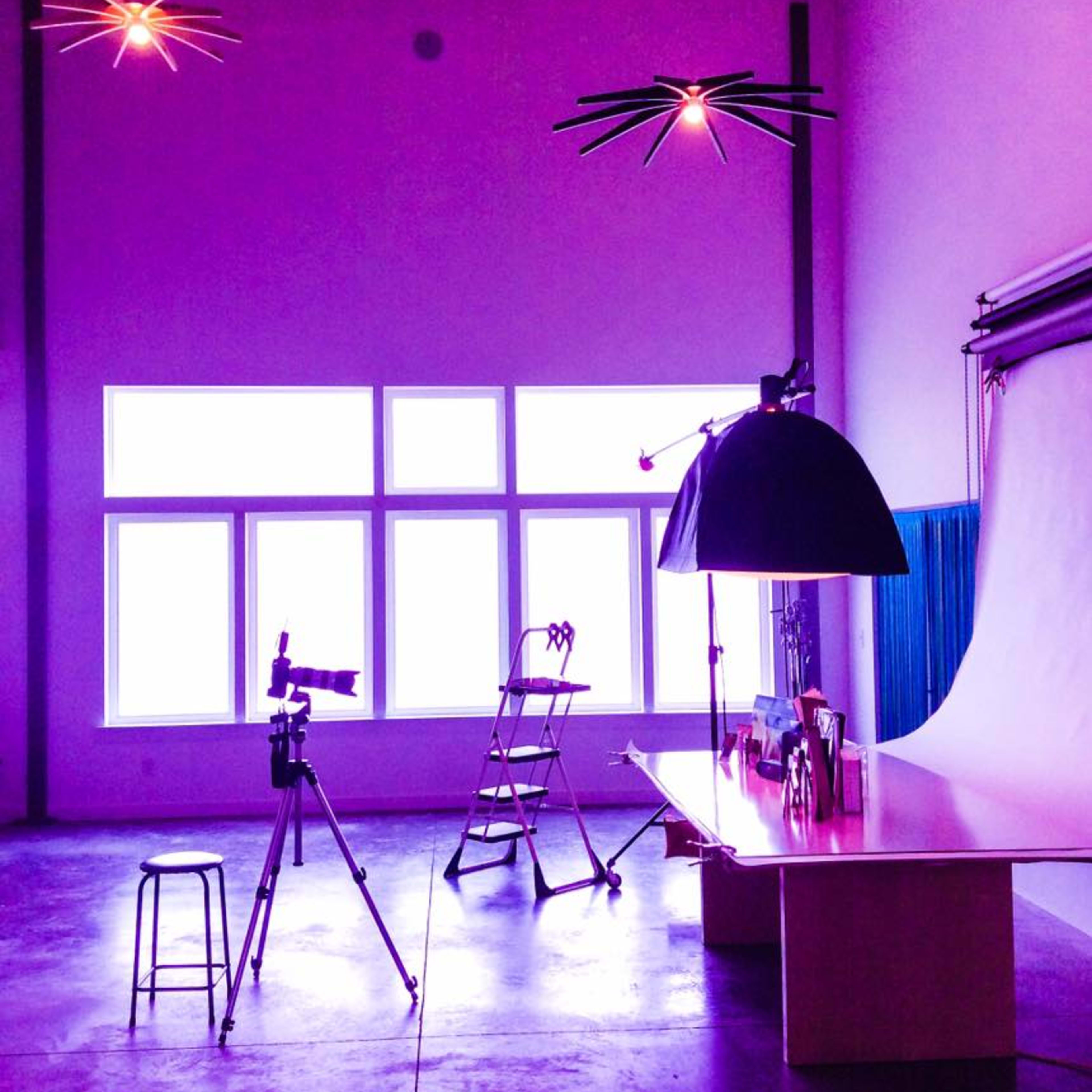 A photography studio features a large backdrop, a stool, a camera on a tripod, and a lighting setup under purple-hued ceiling lights.