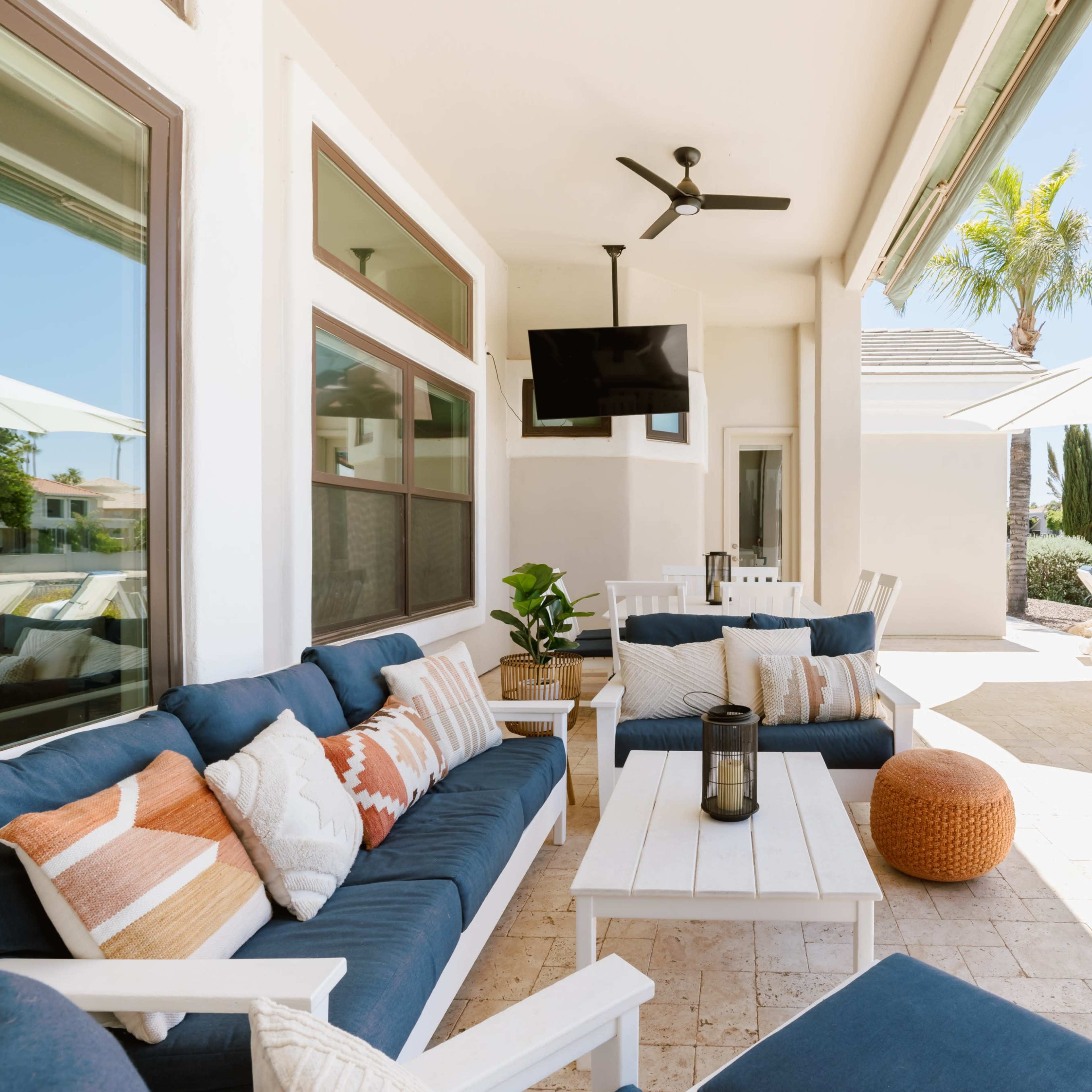 A patio area features a sectional sofa, a coffee table, a lantern, and a television, with lounge chairs and umbrellas visible nearby.