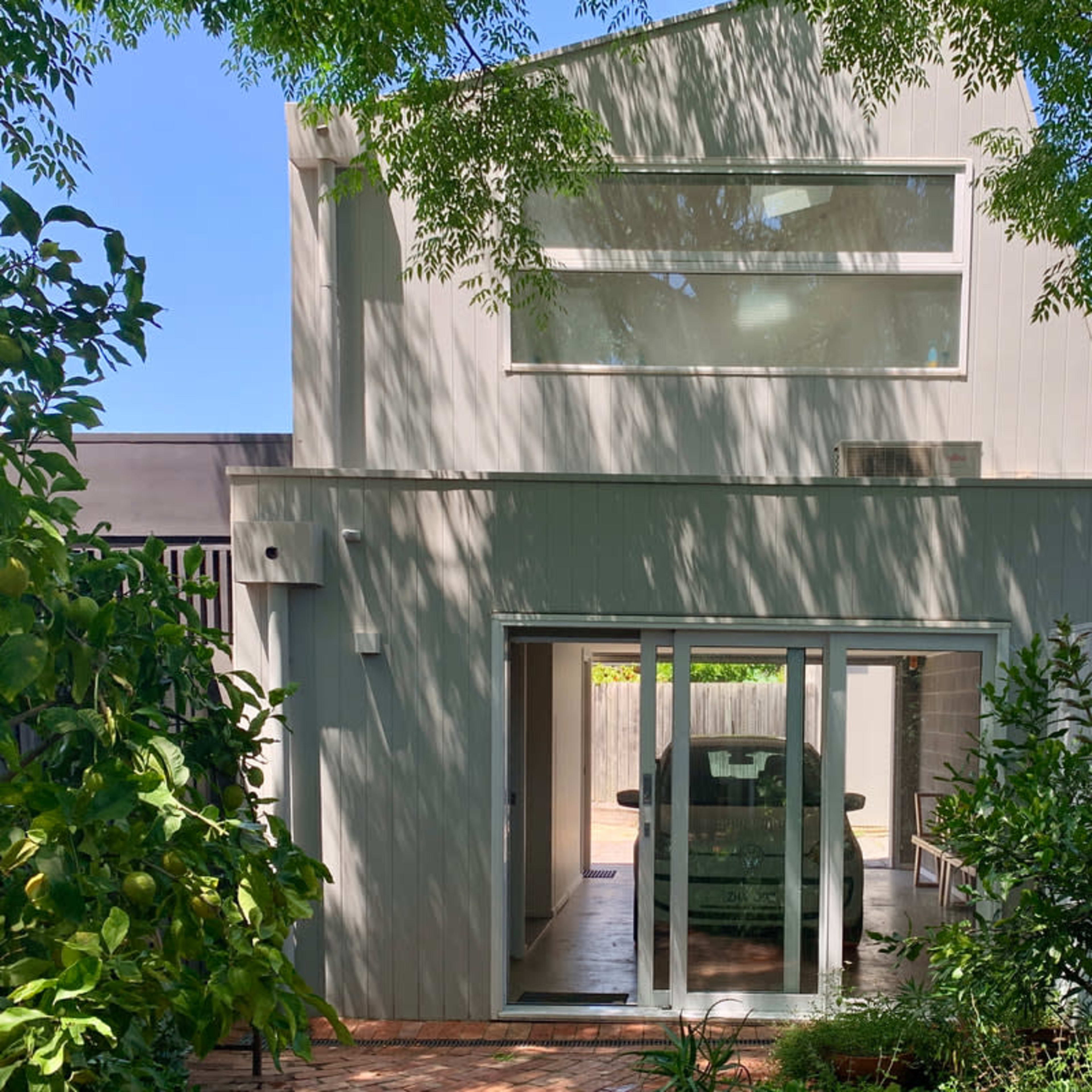 A two-story house with large windows and a car visible through the sliding glass doors is surrounded by greenery.