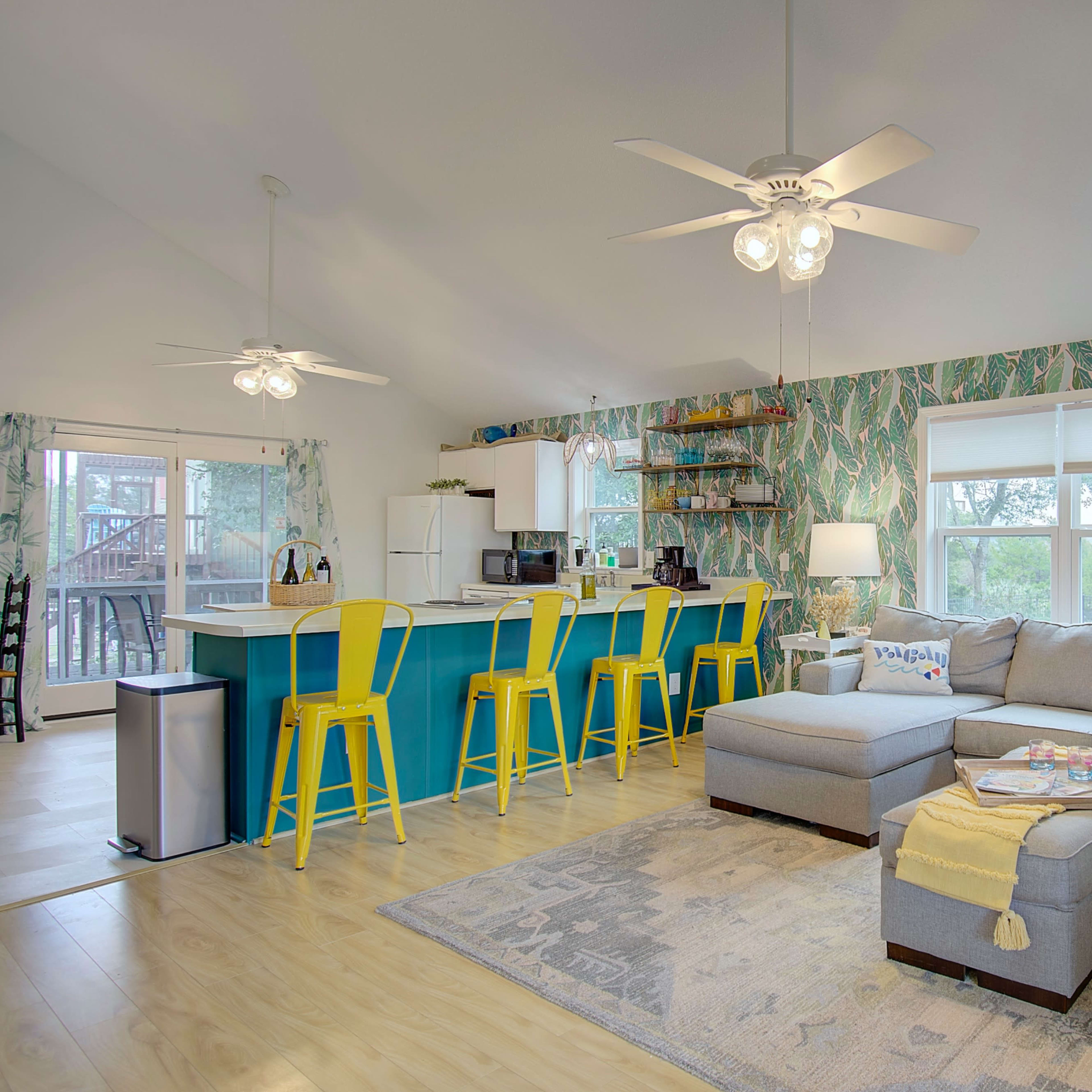 The image shows a bright, open living space with a sectional sofa, a kitchen island with yellow stools, and tropical-themed wallpaper.