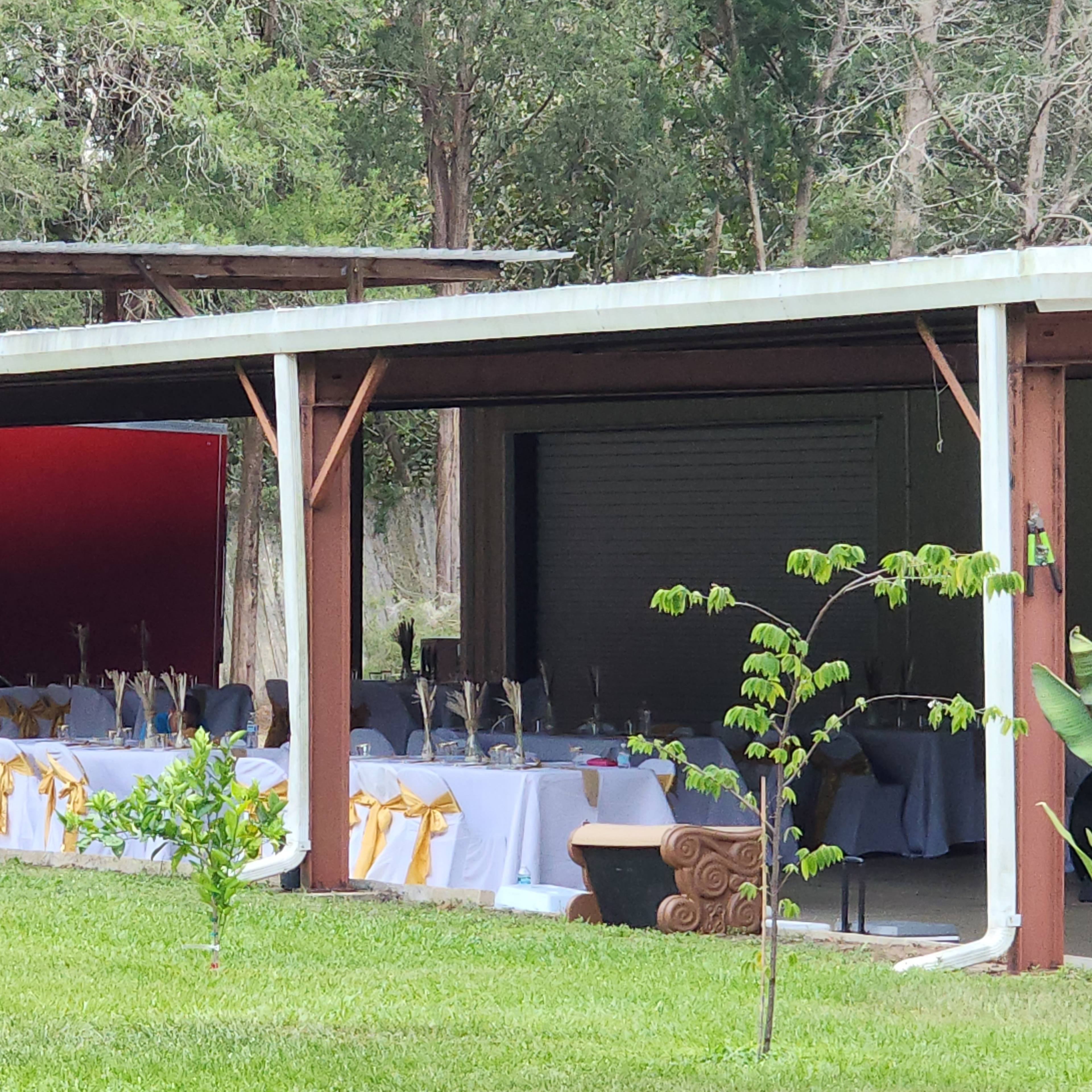 A covered outdoor area displays several tables set with white tablecloths and gold sashes, surrounded by greenery.