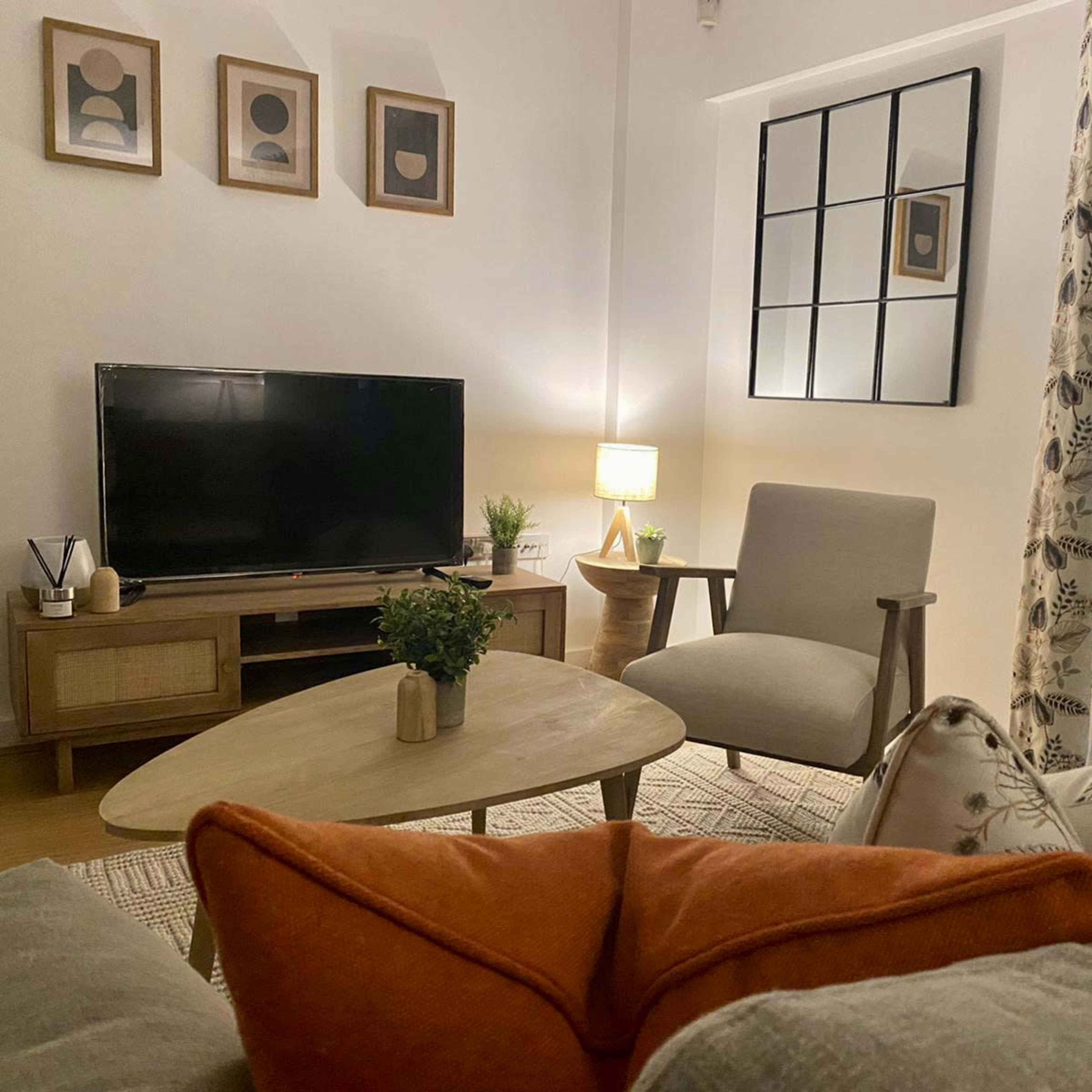 A cozy living room features a television, a wooden coffee table, an armchair, and decorative pillows on a sofa, with plants and artwork on the walls.