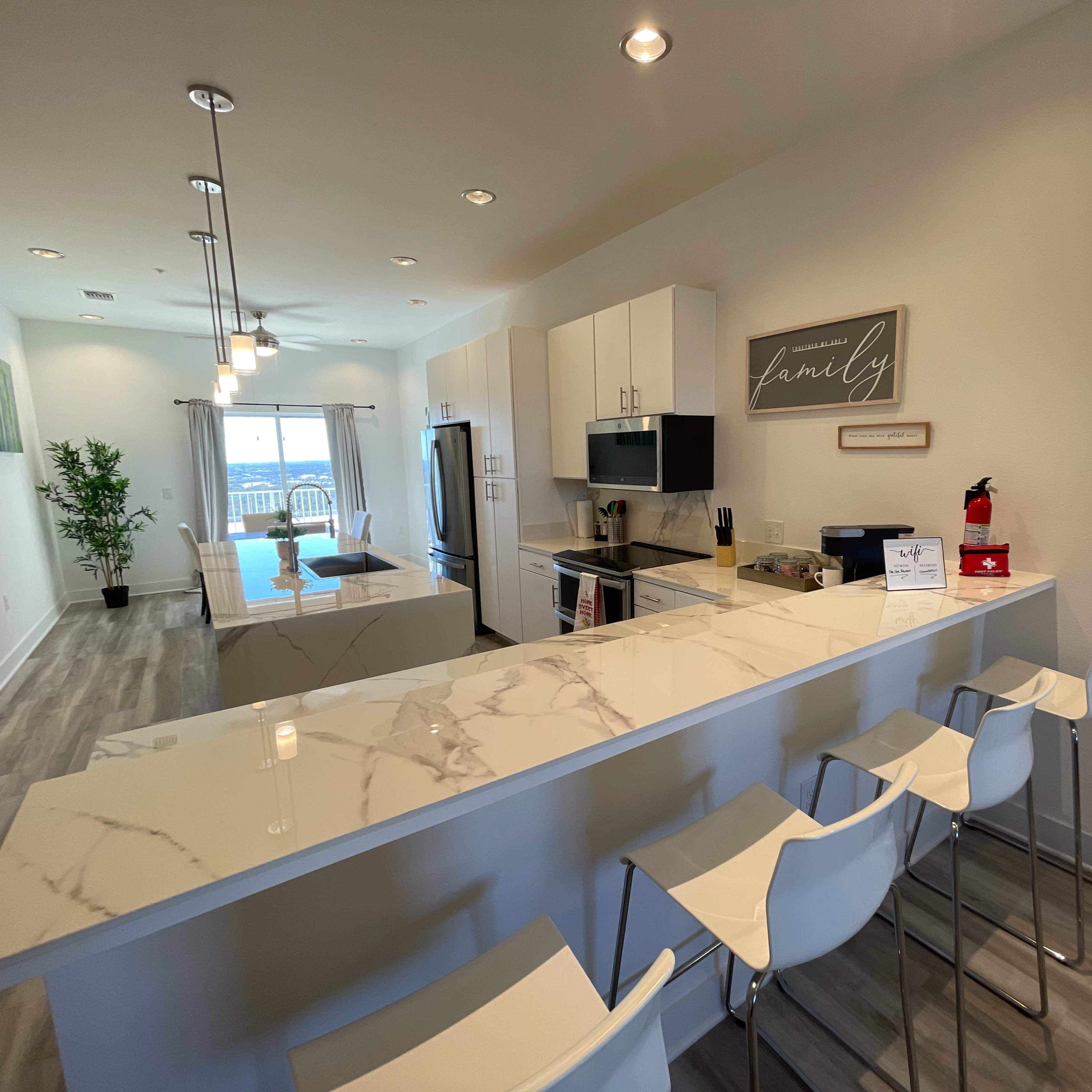 A modern kitchen with white cabinetry, a marble countertop, and bar stools, leading into a living area with a view of a balcony.