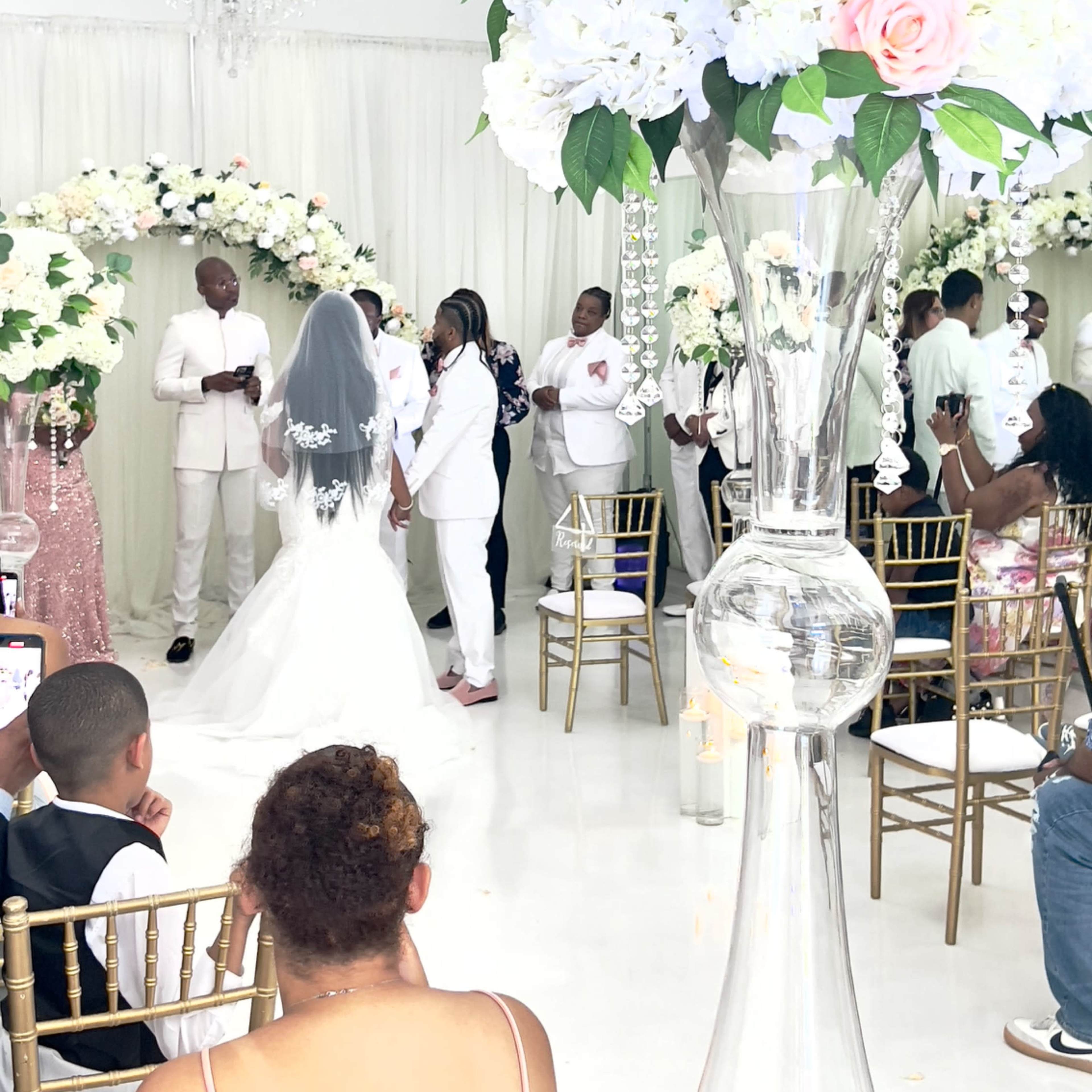 A wedding ceremony is taking place in a decorated venue with floral arrangements, where the couple stands at the altar surrounded by guests.