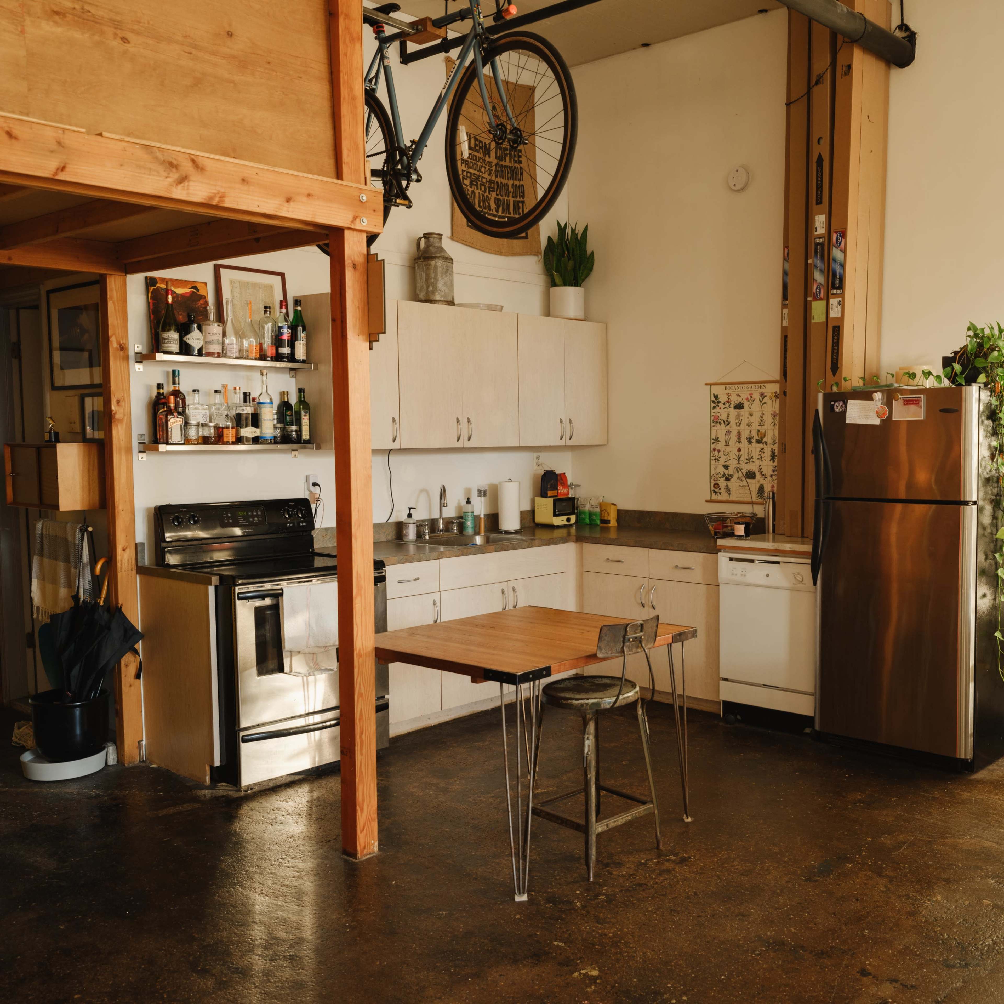 A compact kitchen and dining area features white cabinets, a stainless steel refrigerator, and a wooden table with metal chairs, while a bicycle hangs from the ceiling.