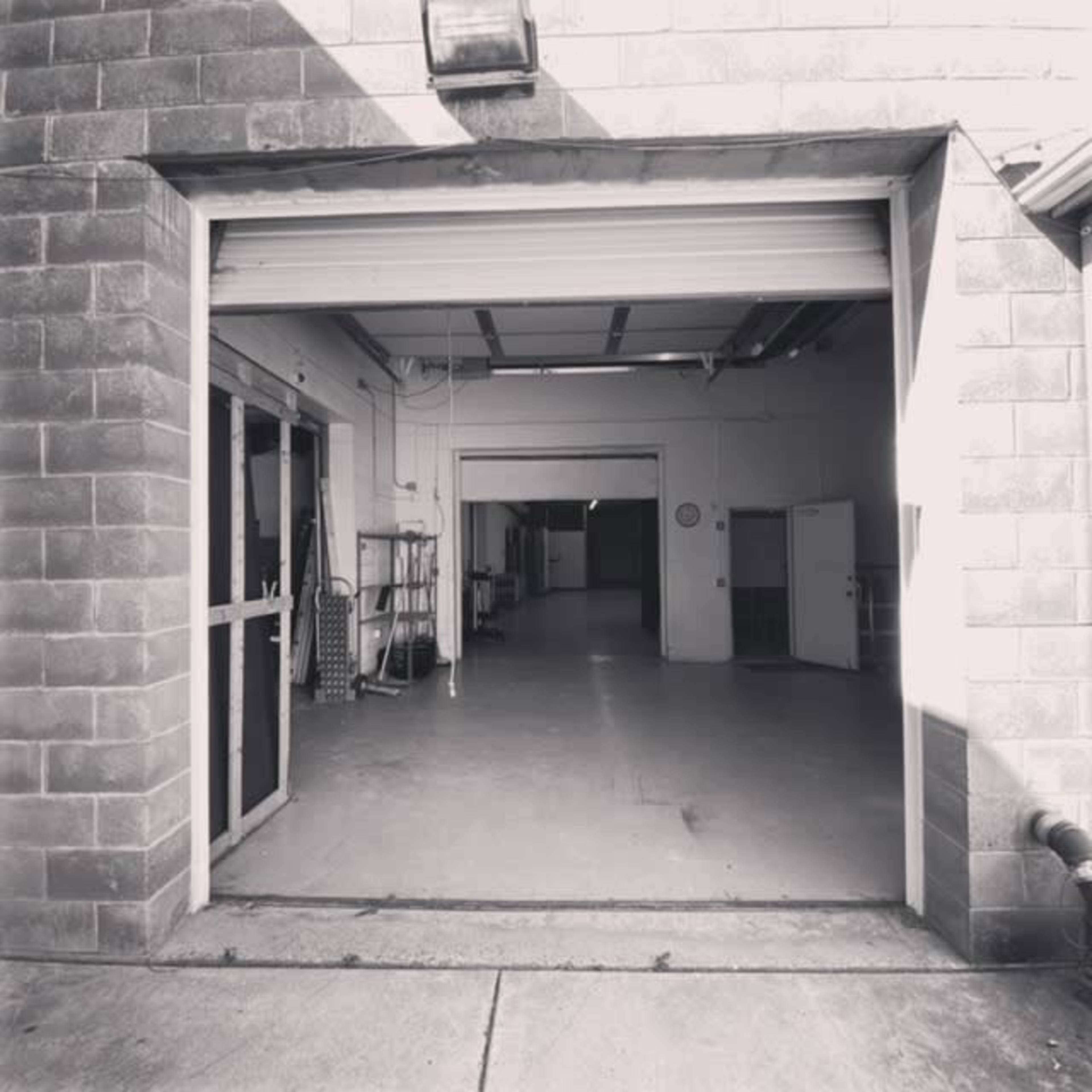 An open garage door leading into a large, empty industrial space with concrete flooring and several doors on the walls.