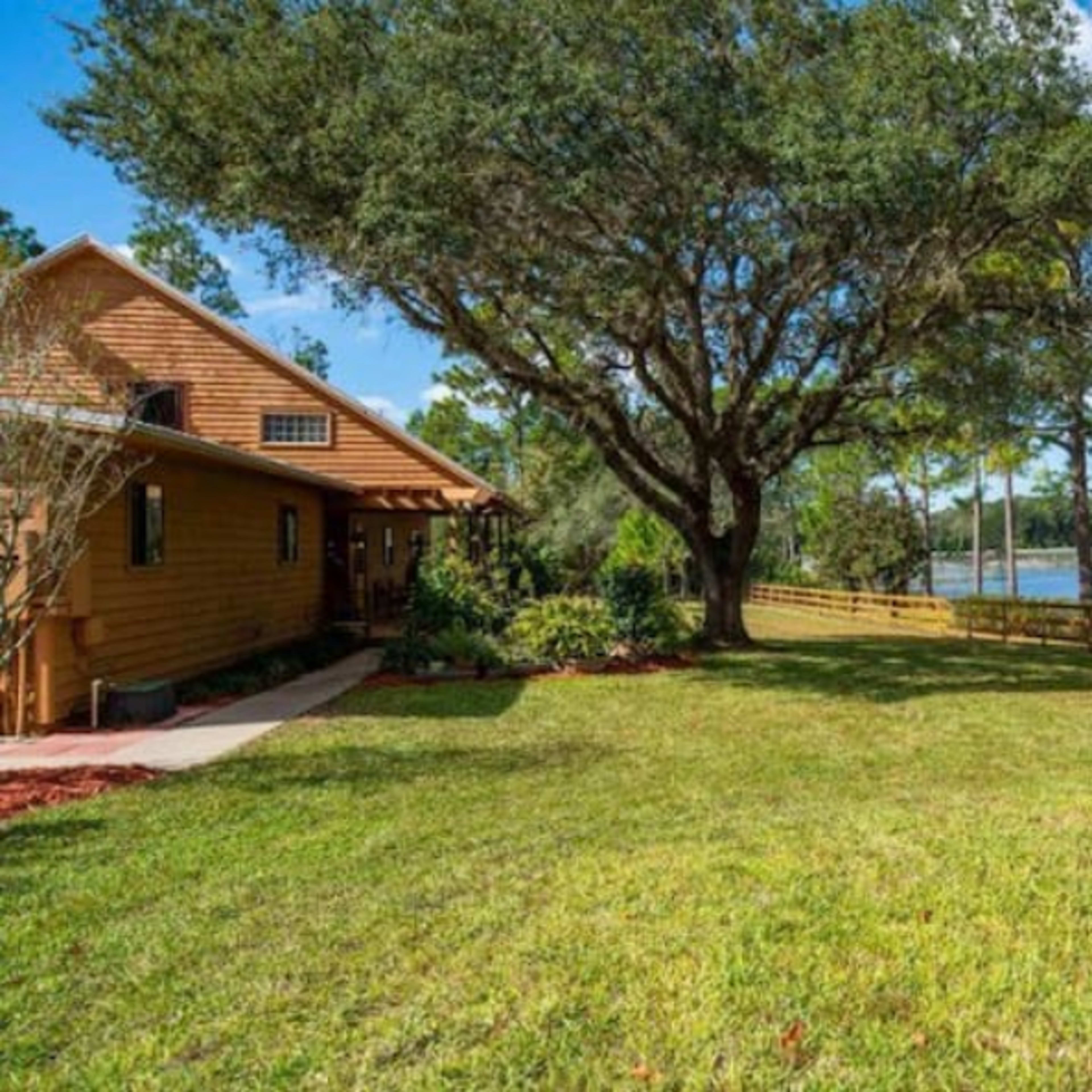 A wooden house with a grassy yard is located near a lake, surrounded by trees and a wooden fence.