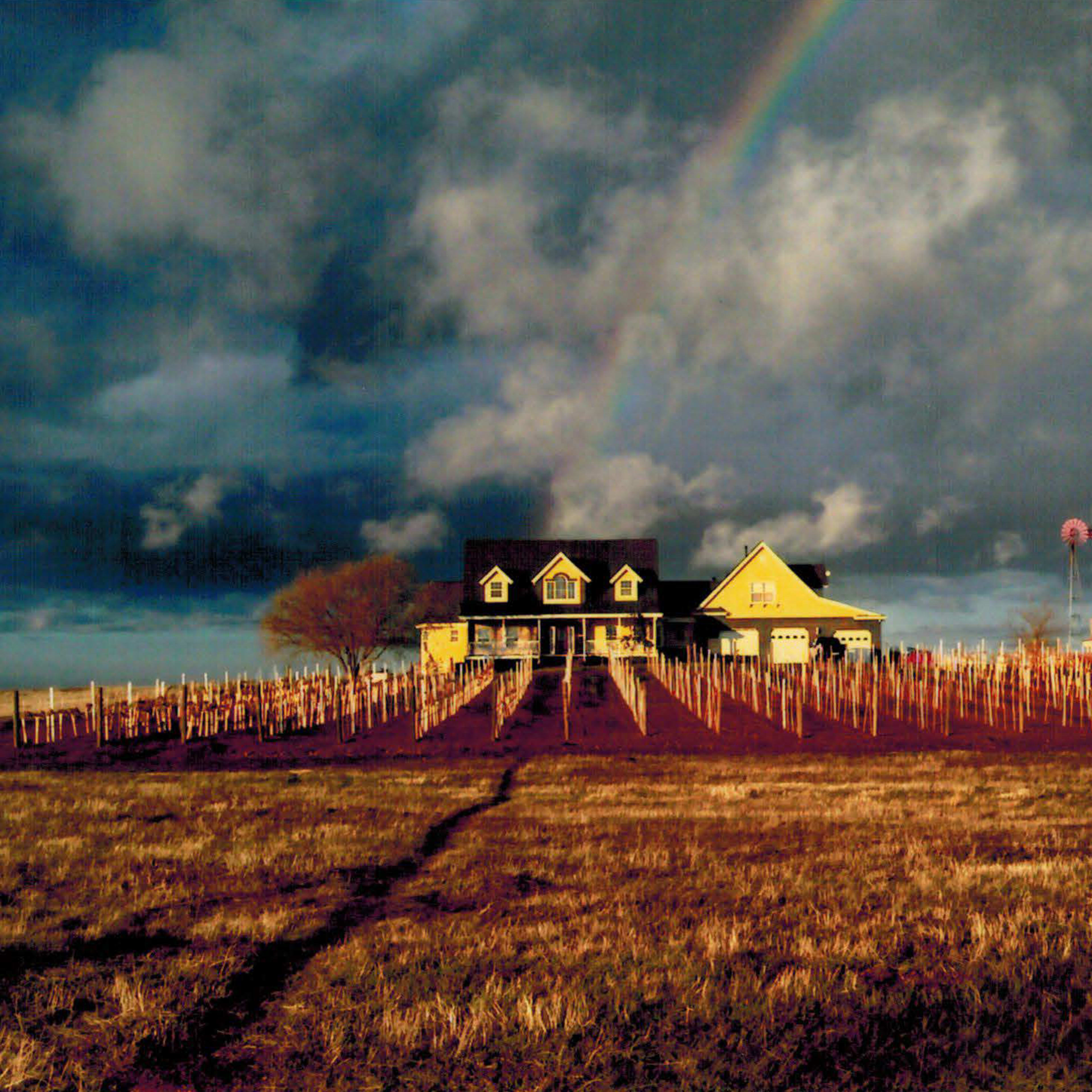 A two-story house with a porch stands on a hill surrounded by a vineyard and a windmill in the background under a partly cloudy sky with a rainbow.