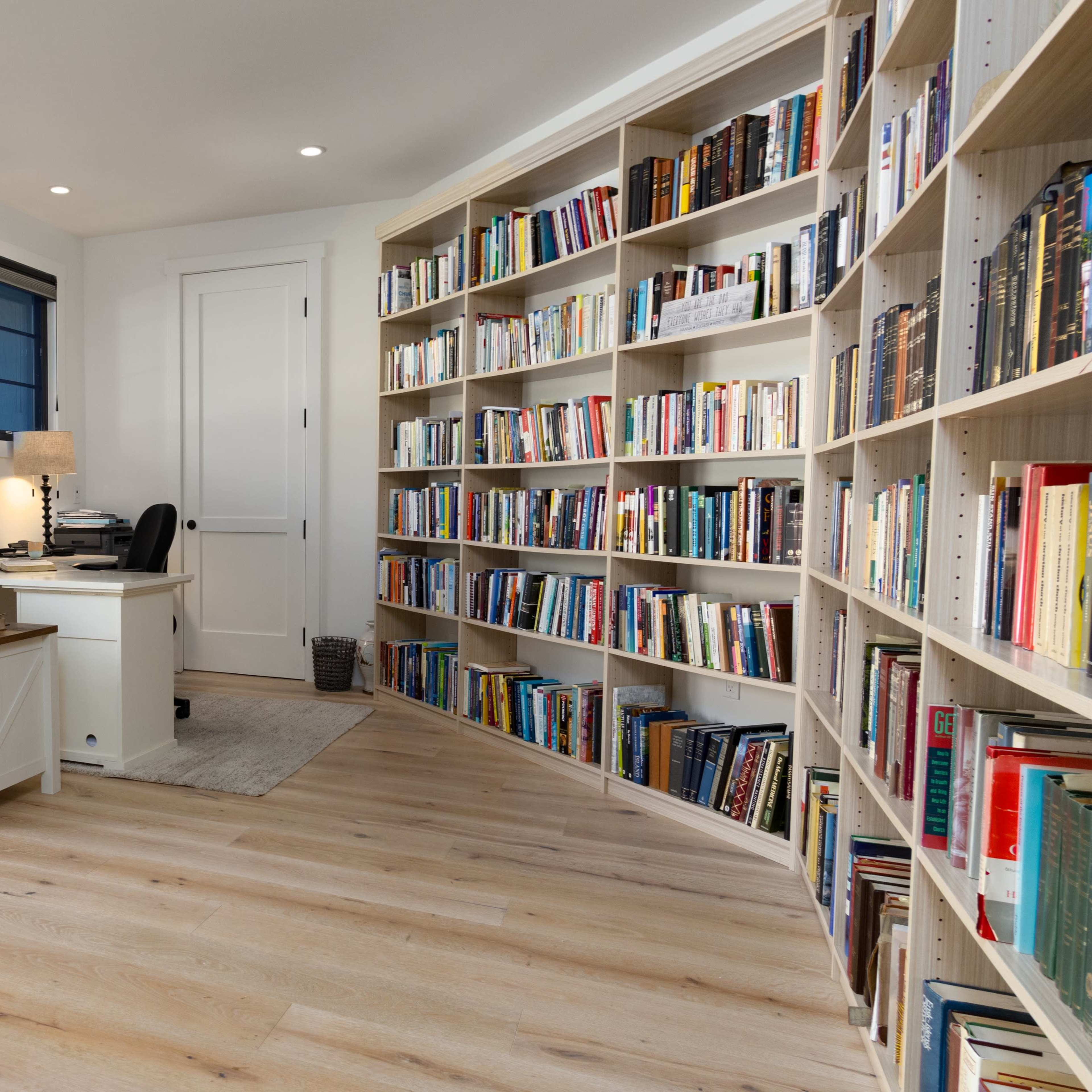 A well-organized home office features a white desk and chair beside a large bookshelf filled with books.
