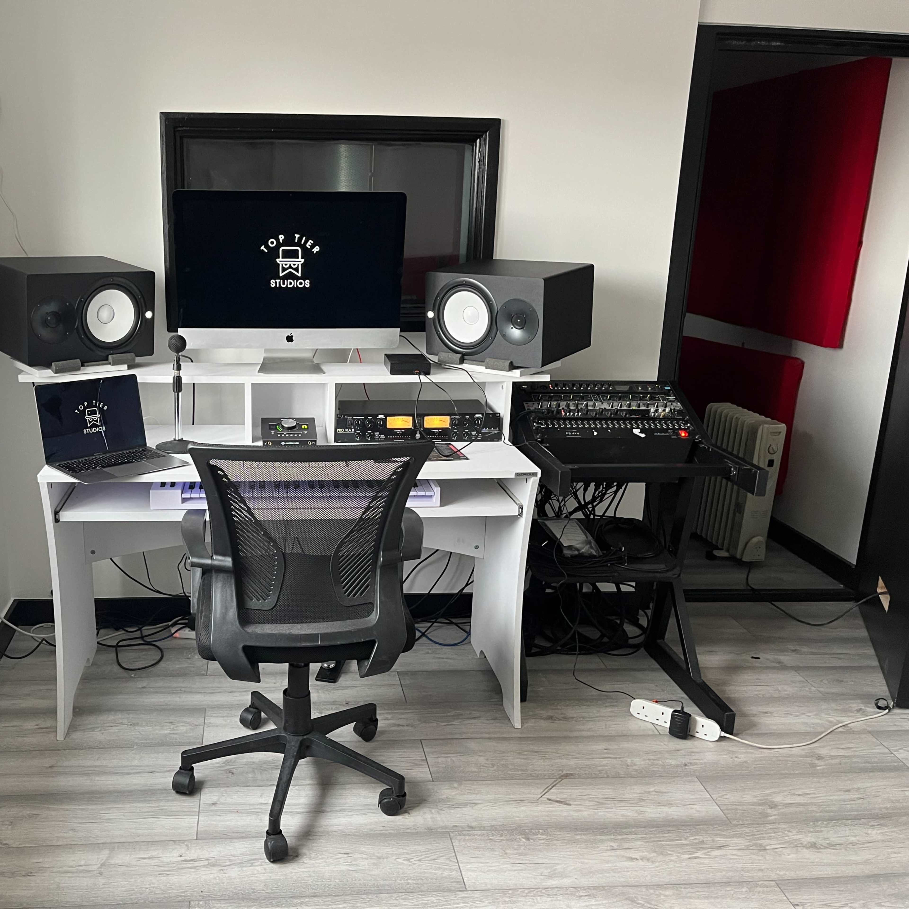 A modern music studio features a desk with computer monitors, audio equipment, and studio monitors positioned in a well-lit room.