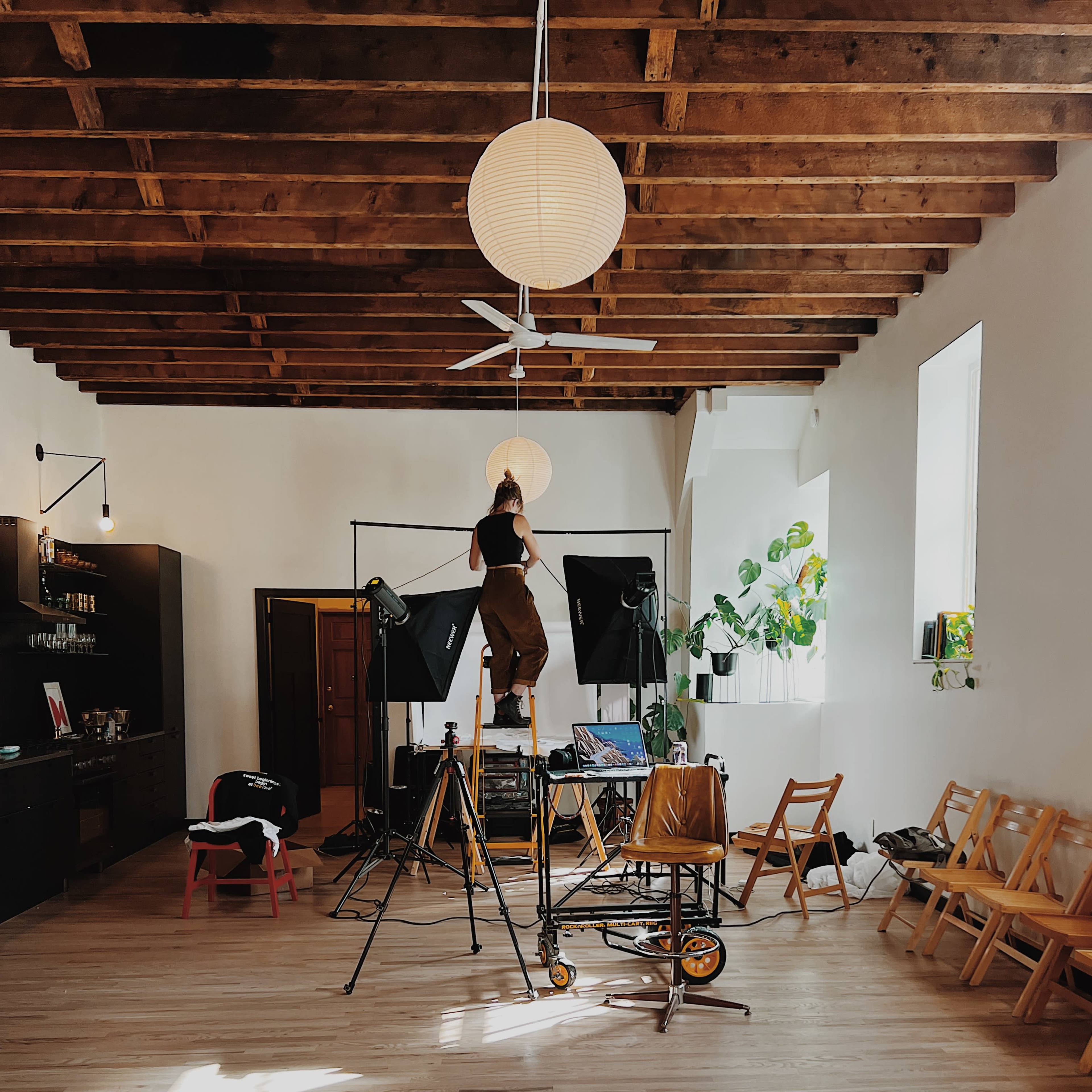 A person stands on a ladder adjusting lights in a spacious studio with wooden beams and minimal furnishings.