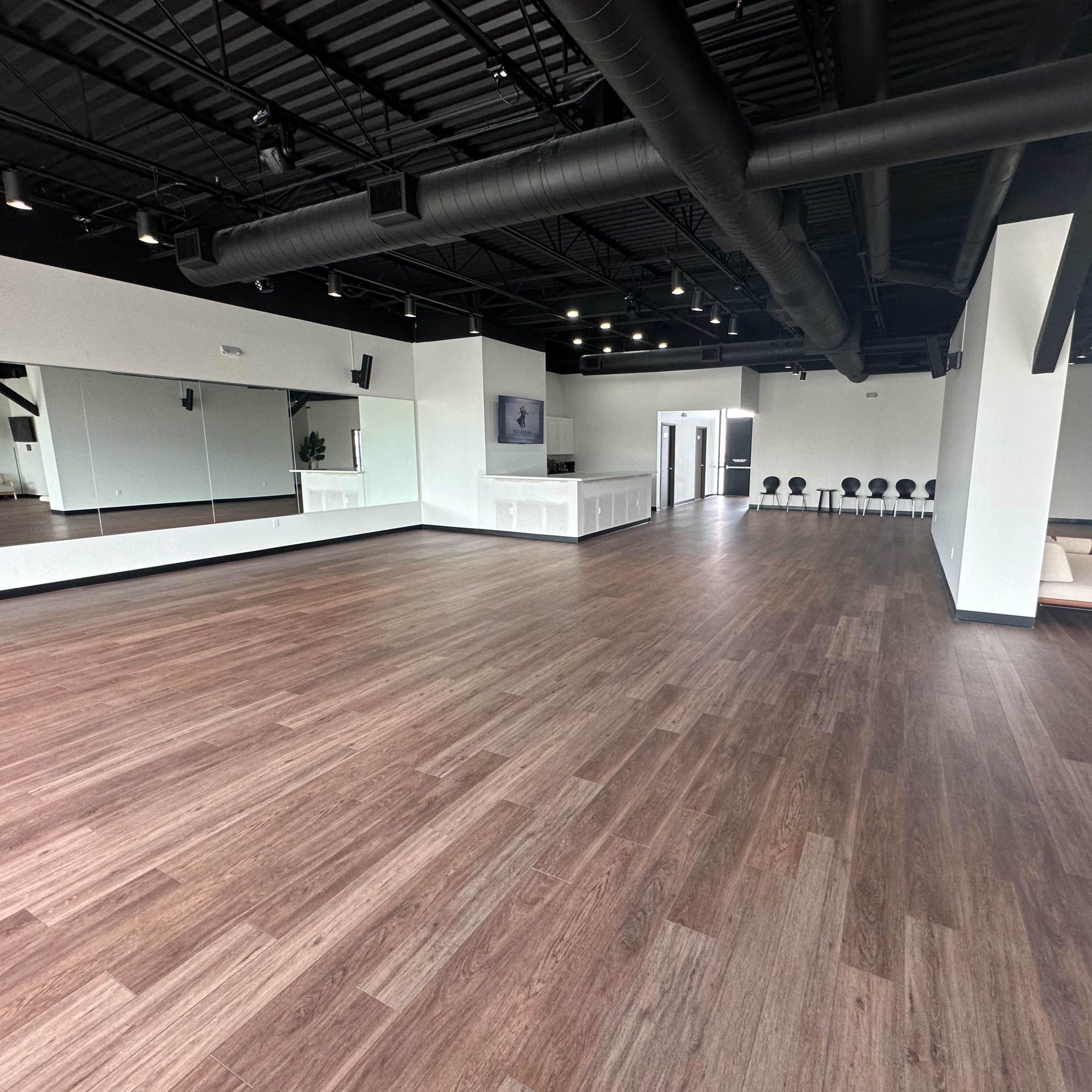 A spacious, well-lit studio with wooden flooring, large mirrors on one wall, and a seating area in the corner.