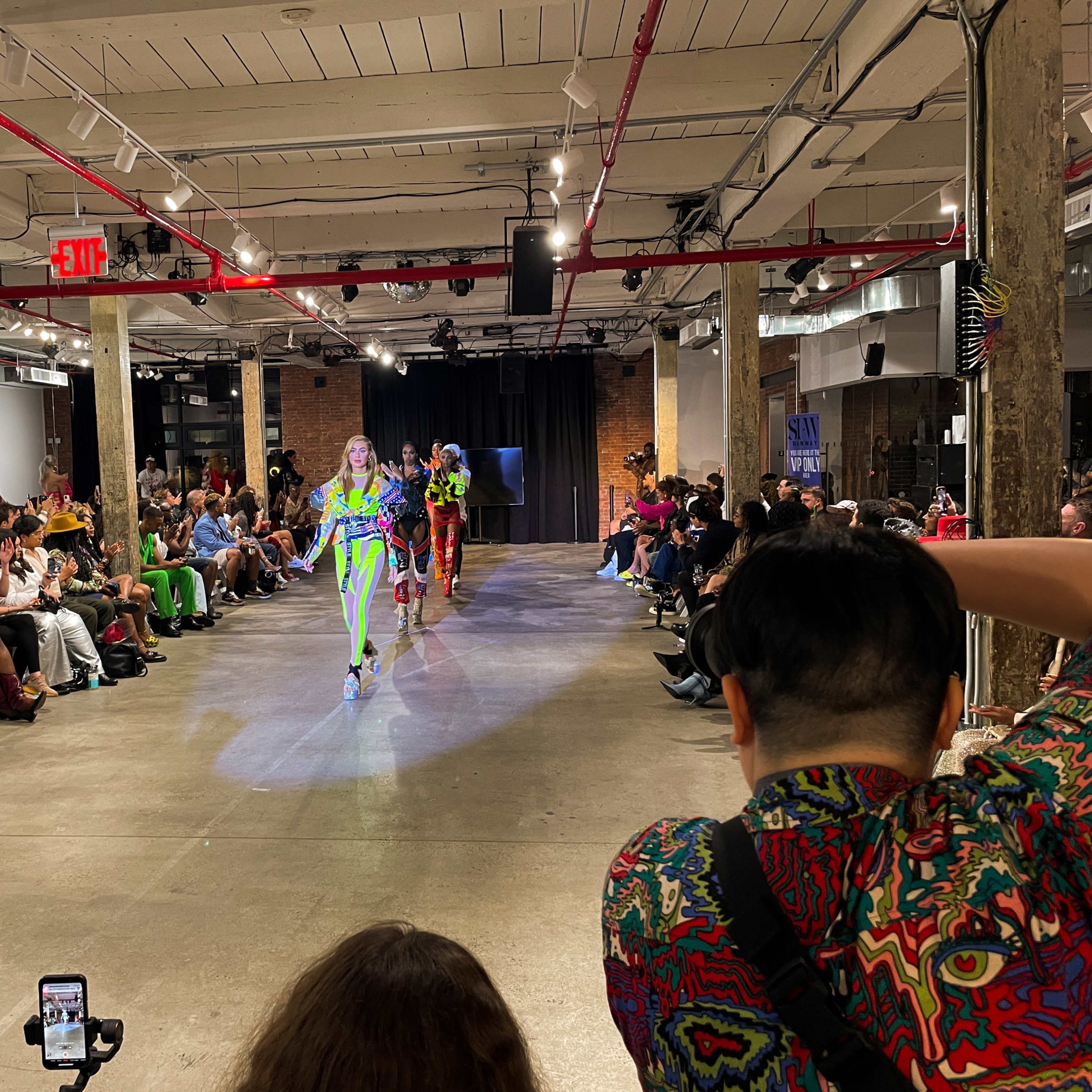 A fashion show is taking place in an industrial space, with models walking down the runway amid a seated audience.