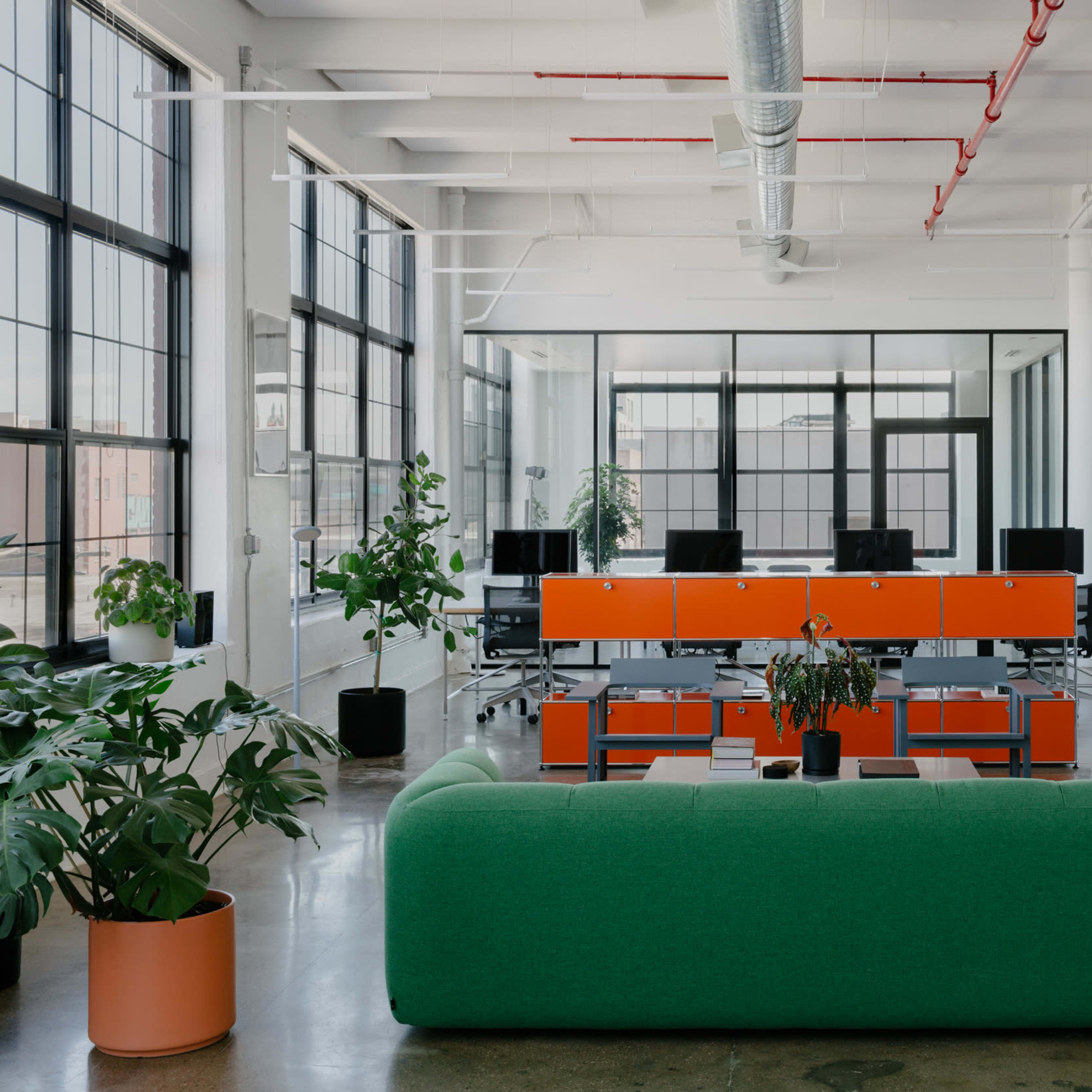 A bright, modern office space features green and orange furniture, large windows, and computer workstations arranged in a collaborative setup.