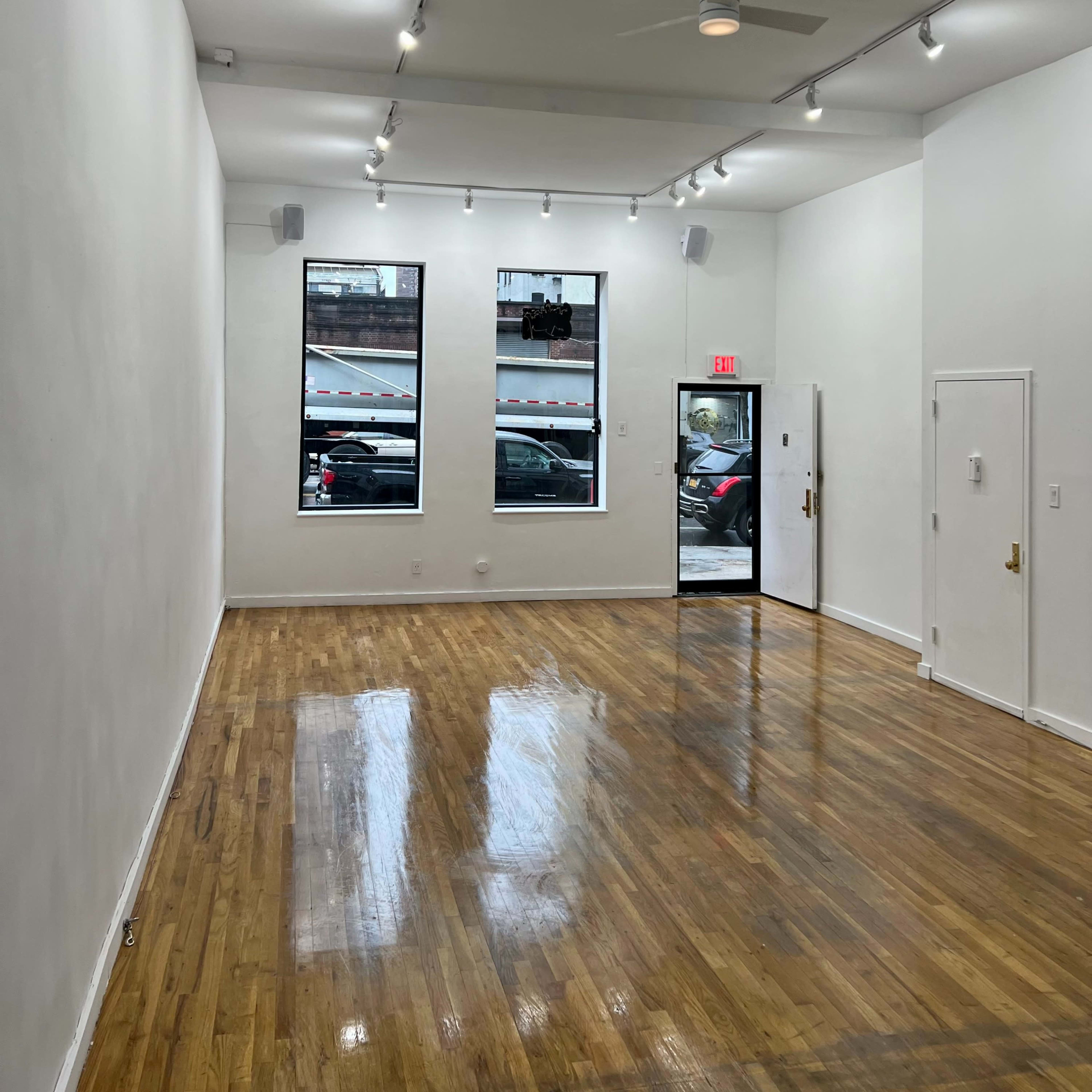 The interior of a spacious, well-lit room with white walls and glossy wooden floors features large windows and a door leading to the street.