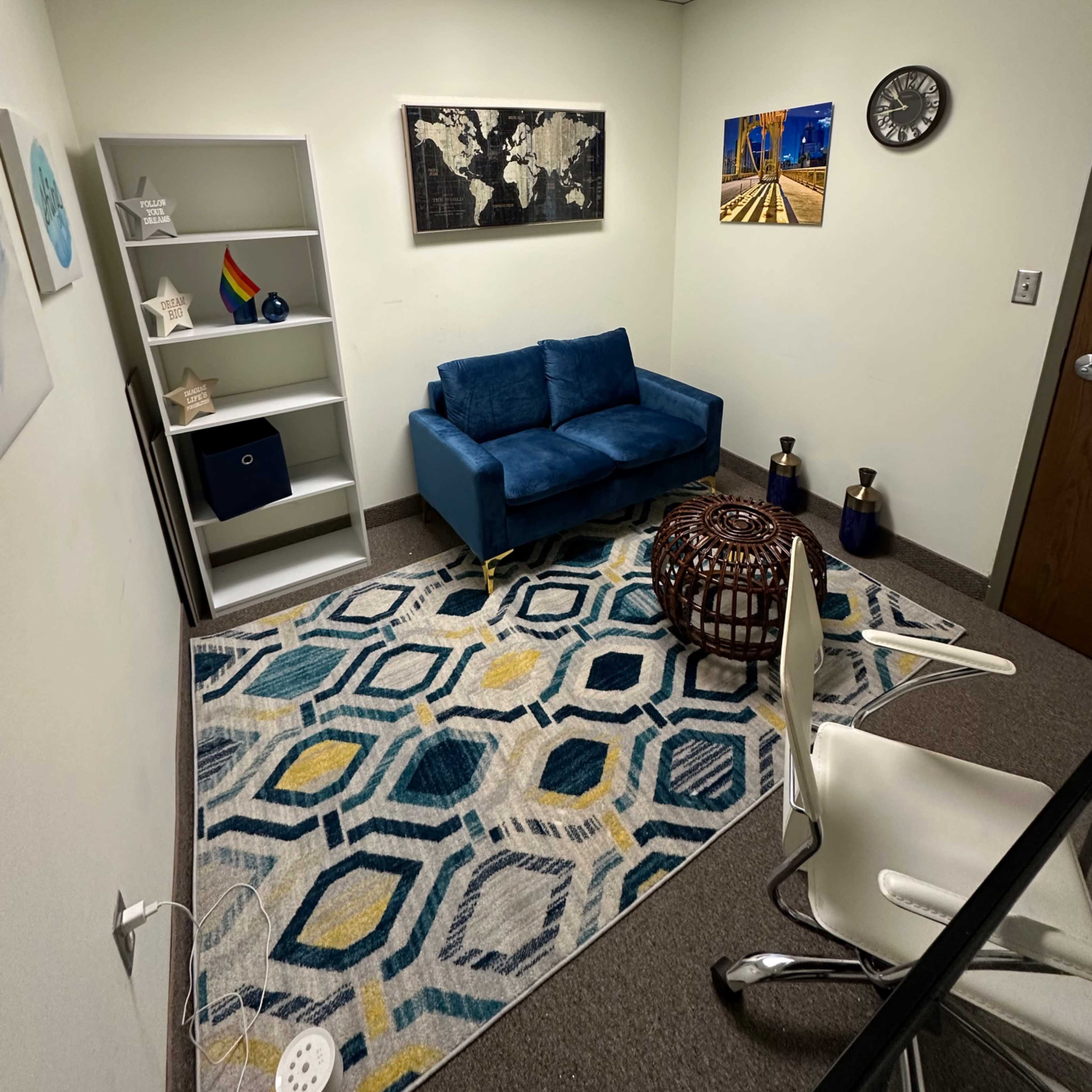 A small office space featuring a blue couch, a decorative rug, a bookshelf, wall art, and a clock.