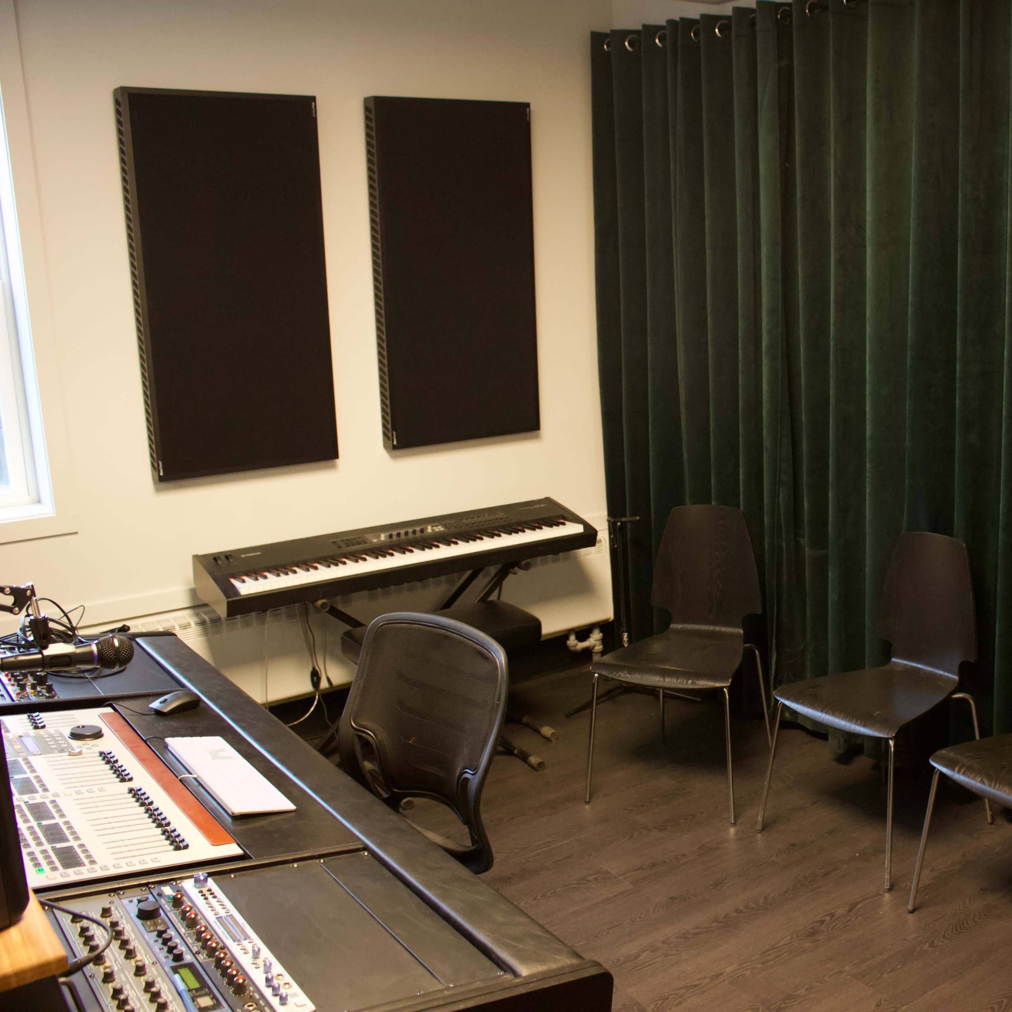A music studio setup with a control desk, a keyboard, and four black chairs arranged in front of green curtains.