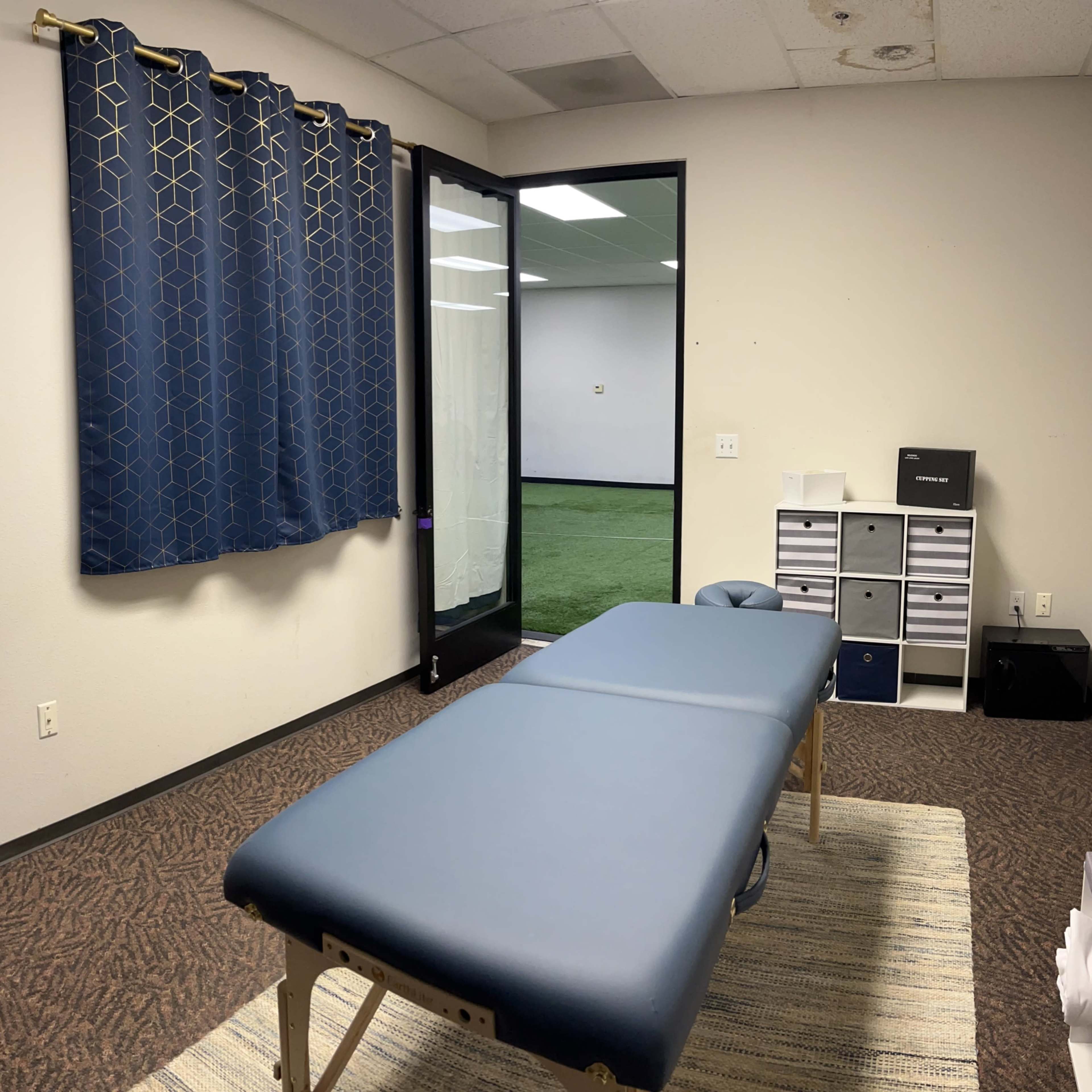 A treatment room with a blue massage table, a doorway leading to another room, and storage units along the wall.