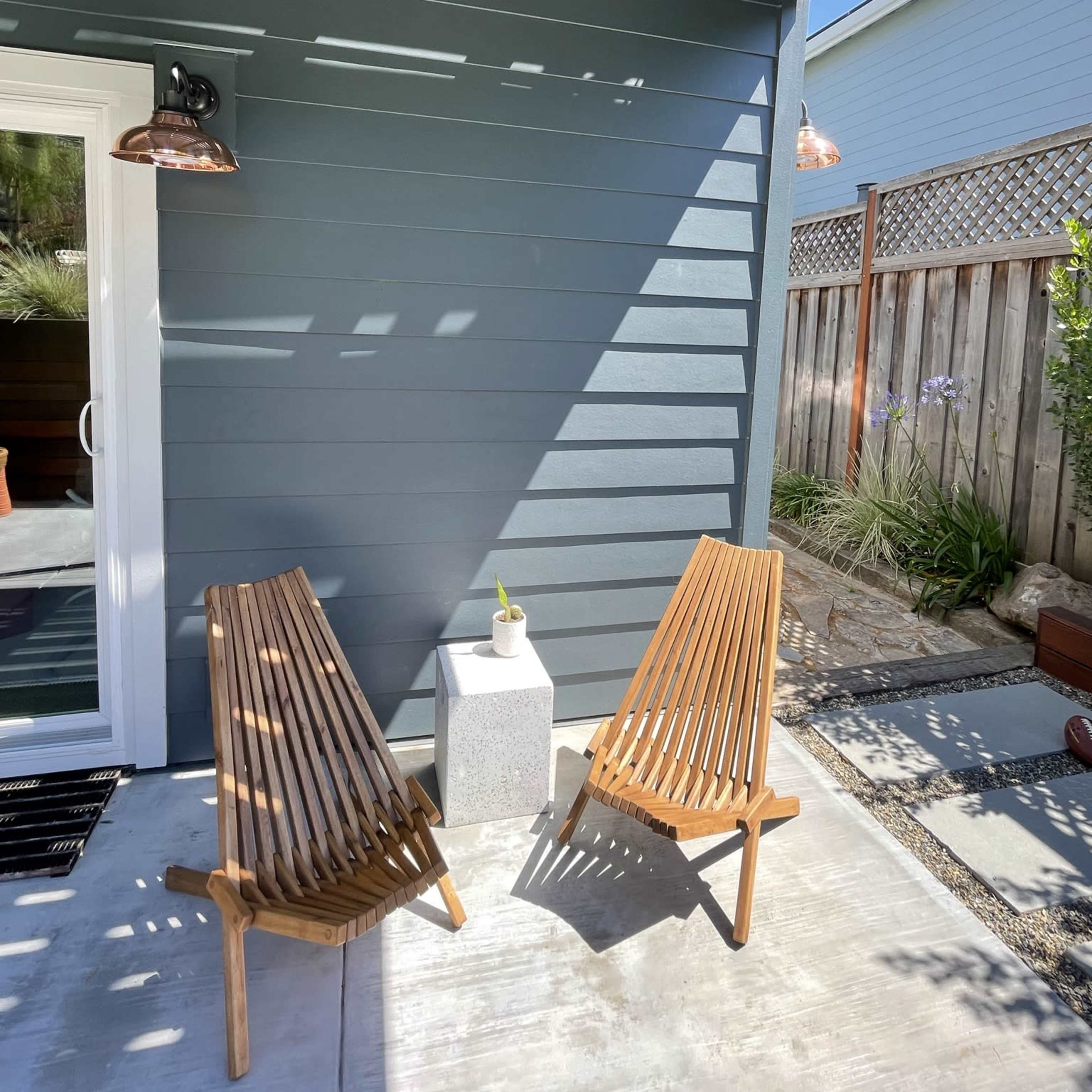 Two wooden lounge chairs beside a small concrete table on a concrete patio next to a gray house wall.