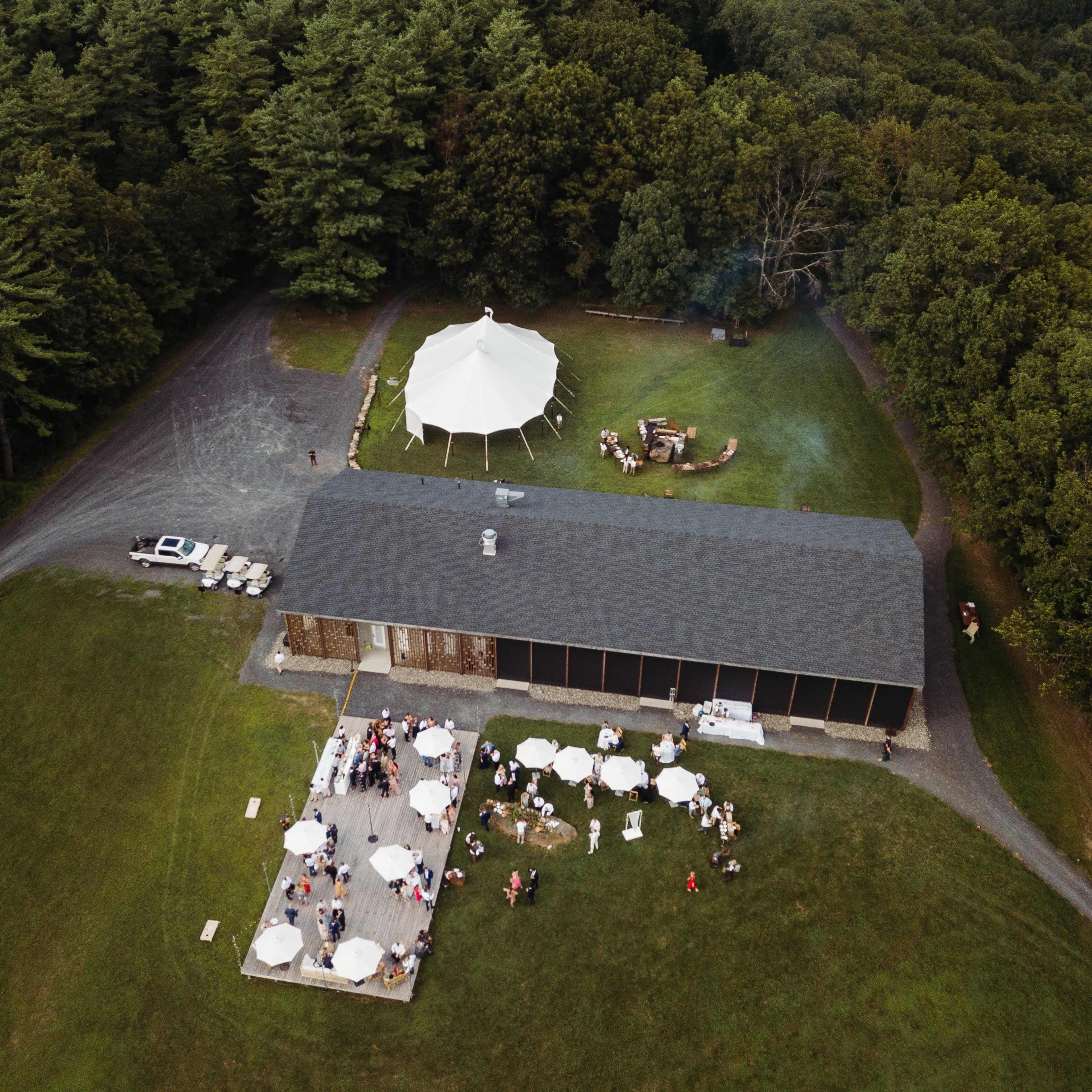 An aerial view shows a large tent set up next to a building, with a gathering of people and tables arranged on a grassy area surrounded by trees.