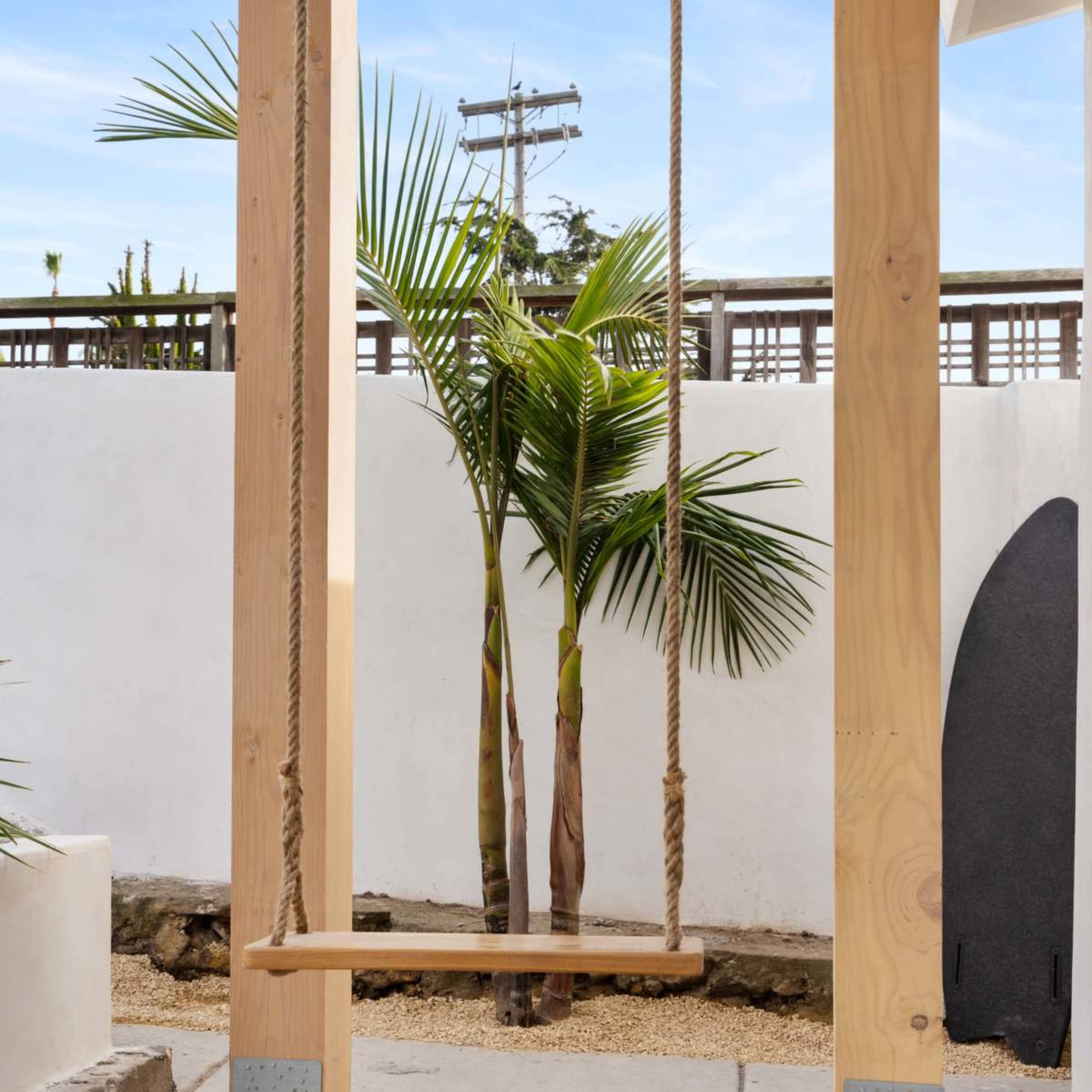 A wooden swing hangs from a frame near two palm trees in a patio area with a concrete floor.