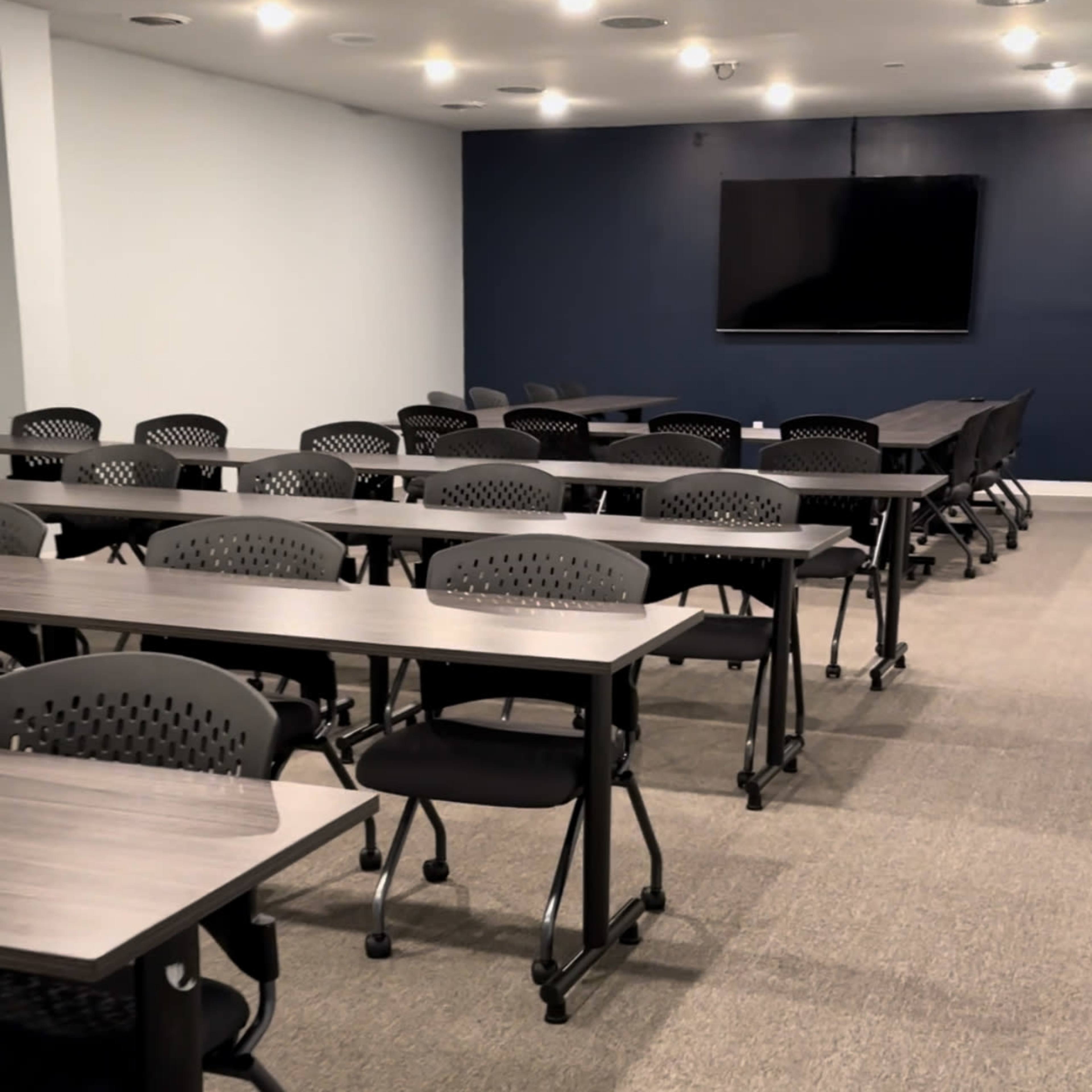 A training room features multiple rows of tables and chairs facing a wall-mounted television.