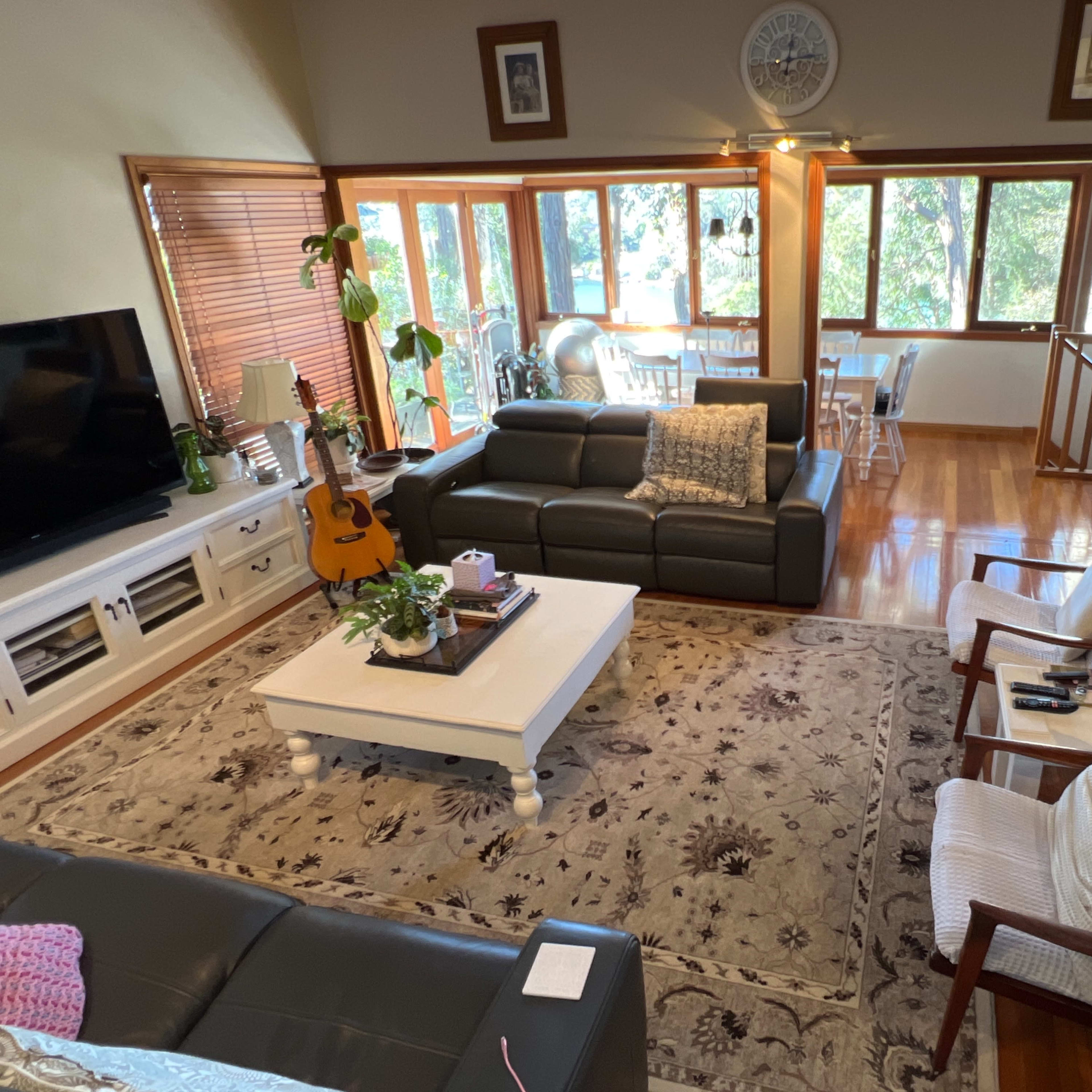A spacious living area with a dark leather sofa, a white coffee table, an acoustic guitar, and large windows overlooking a natural setting.