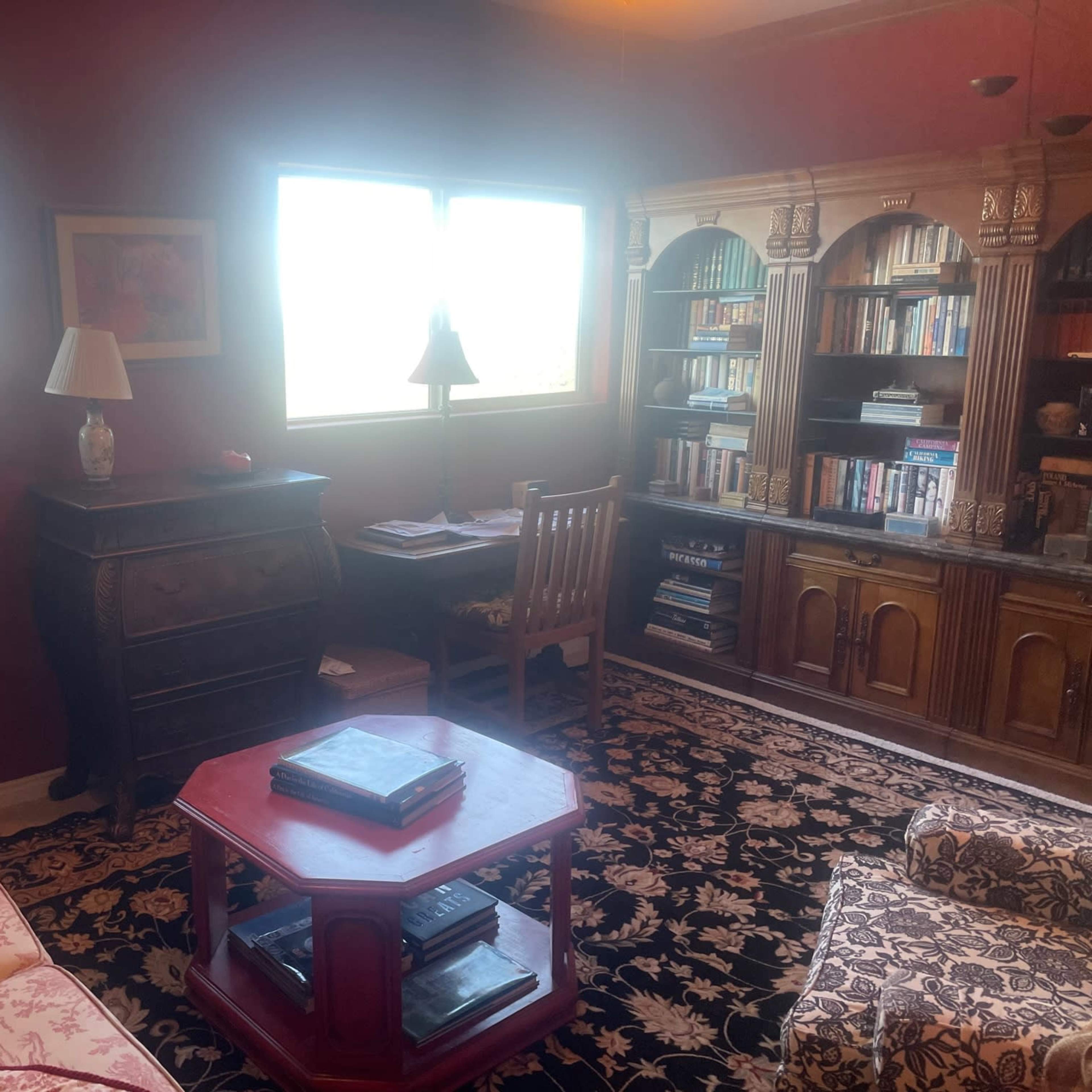 A cozy living room with a floral patterned couch, a wooden desk, a bookshelf filled with books, and windows that let in natural light.